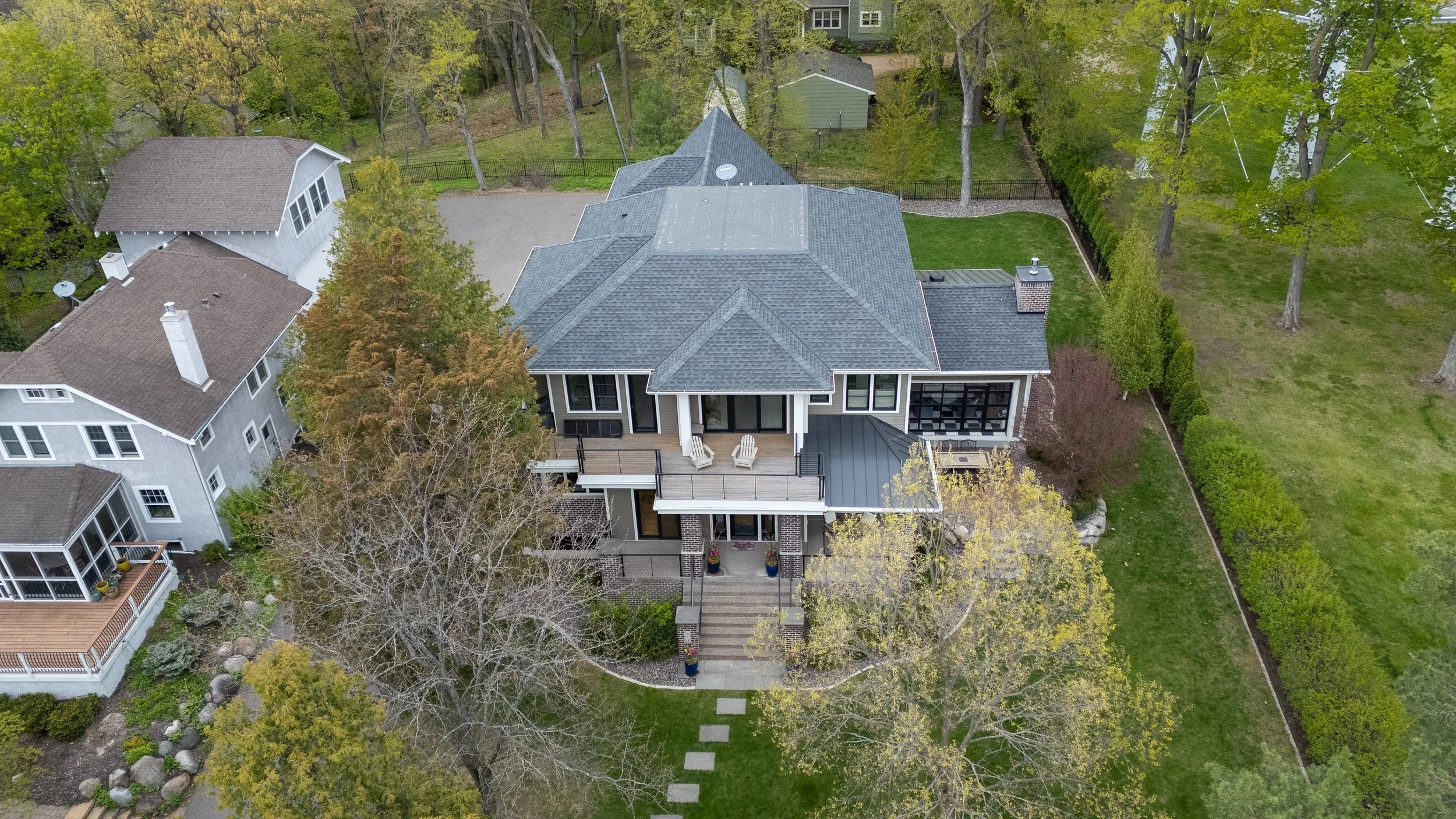 An aerial view of a suburban house with a gray roof, large deck with two chairs, surrounded by green trees, neighboring houses, and a grassy backyard.