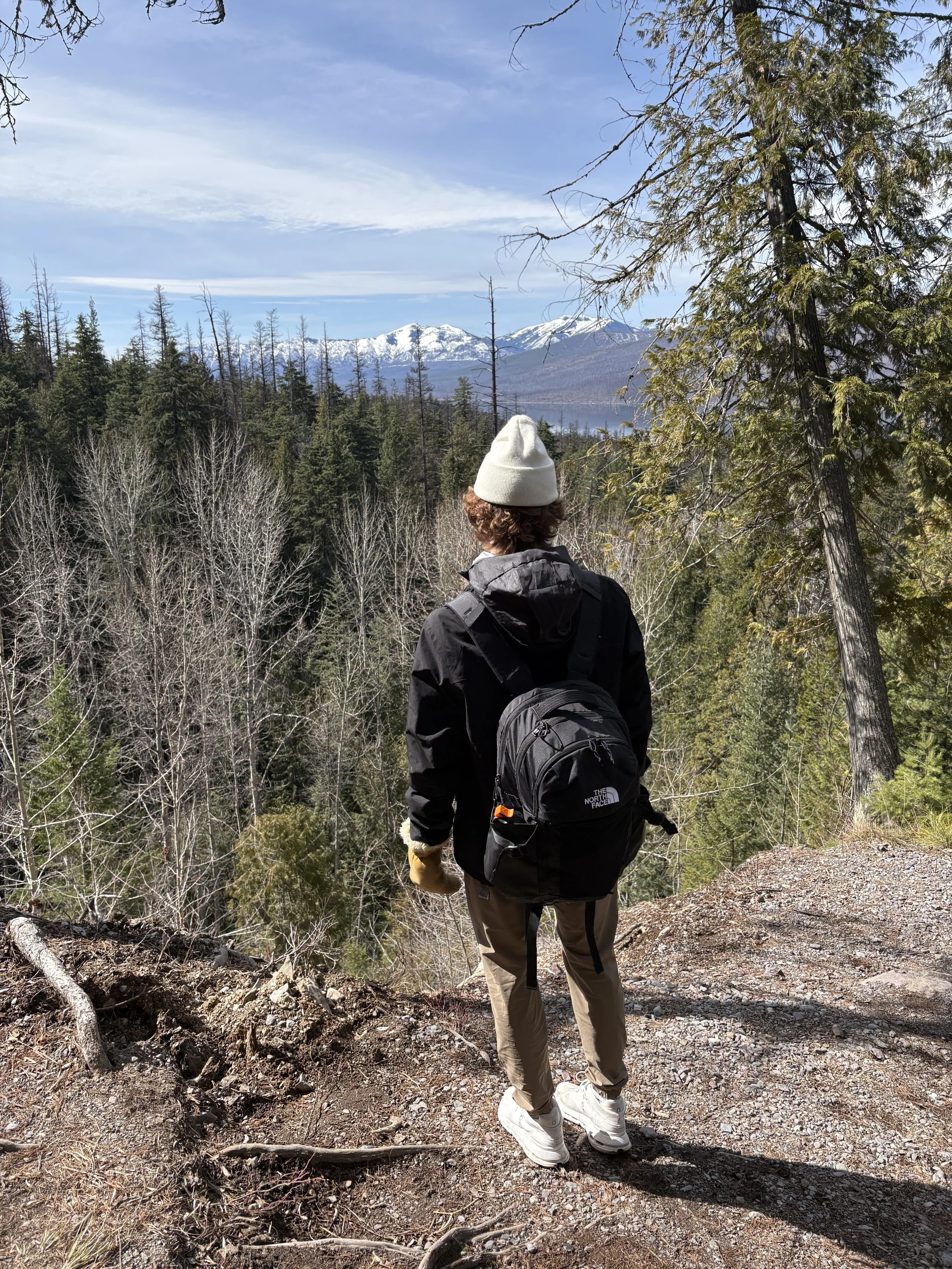 A person with curly hair, wearing a white beanie, black jacket, tan pants, and white sneakers, walking on a dirt trail through a forest with high trees, some with snow on the mountain peaks in the distance, under a partly cloudy sky.