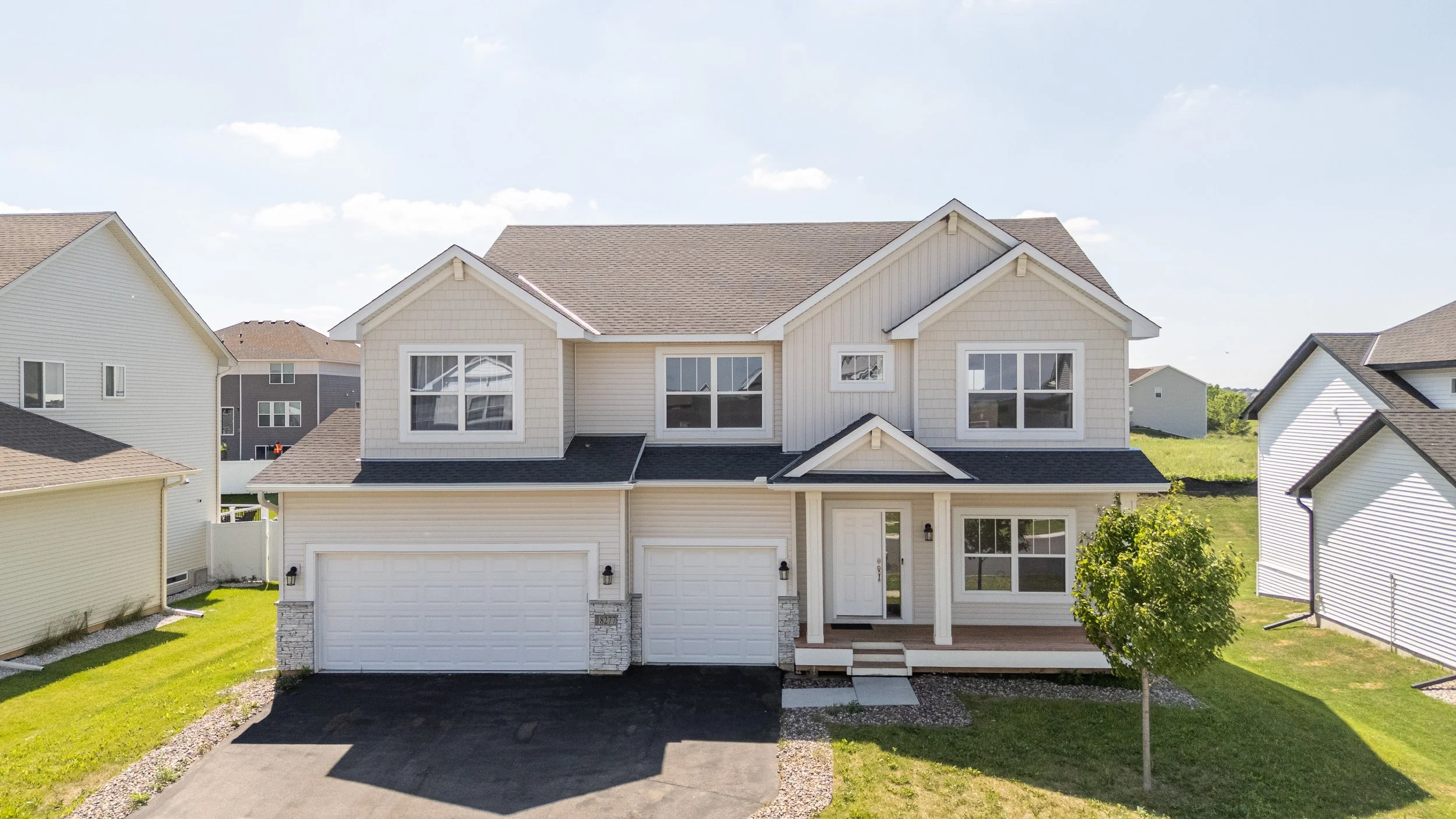 Front view of a two-story, white suburban house with a two-car garage and a small front porch, surrounded by green lawn and neighboring houses under a blue sky with a few clouds.