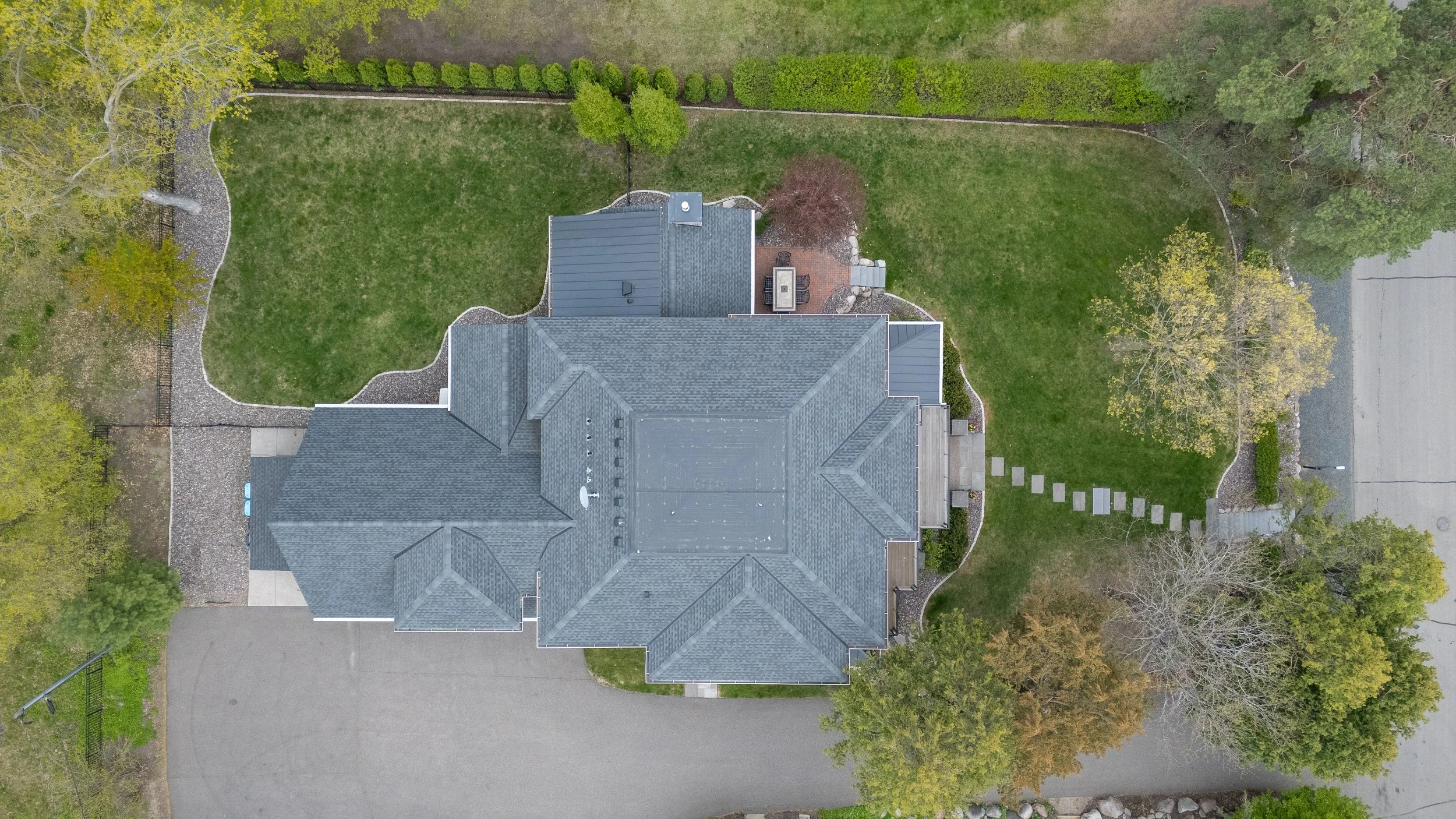Aerial view of a house with gray roof shingles, surrounded by a well-maintained lawn, trees, and a backyard patio area.
