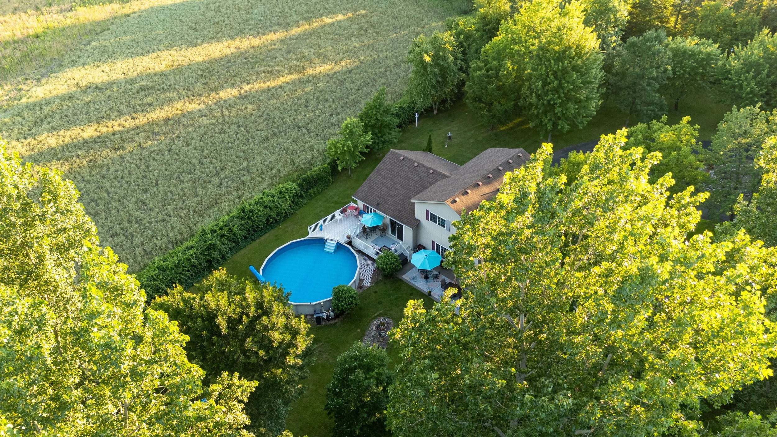 Aerial view of a house with a backyard swimming pool, surrounded by lush green trees and a neighboring farmland.