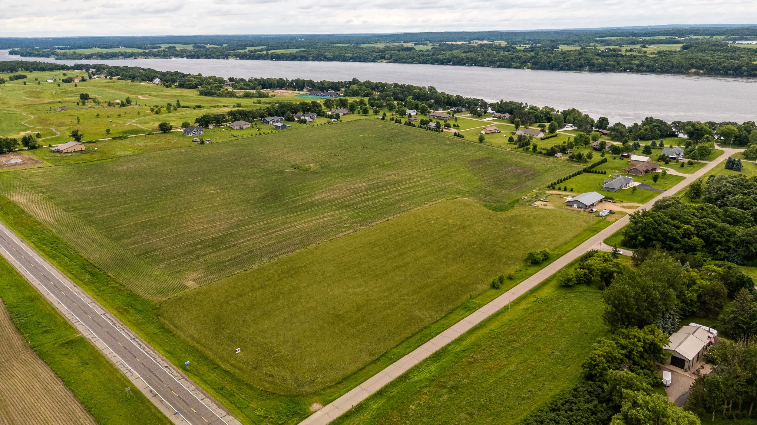 Aerial view of a suburban area near a river, with houses, green lawns, trees, roads, and farmland.