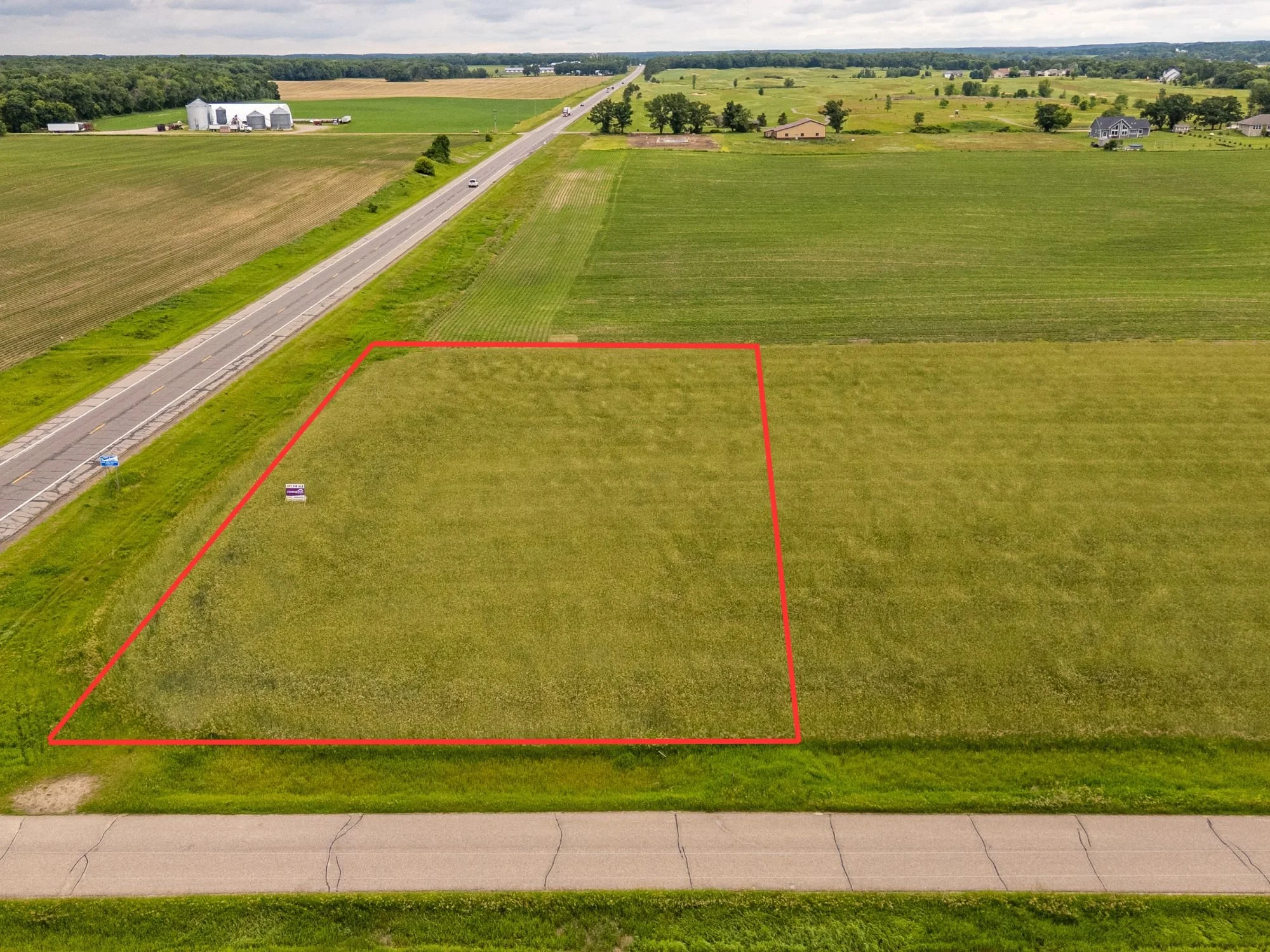 Aerial view of a rural landscape with green farmland, a paved road running through it, and some farm buildings and houses in the background. A red outline marks a rectangular plot of land in the foreground.