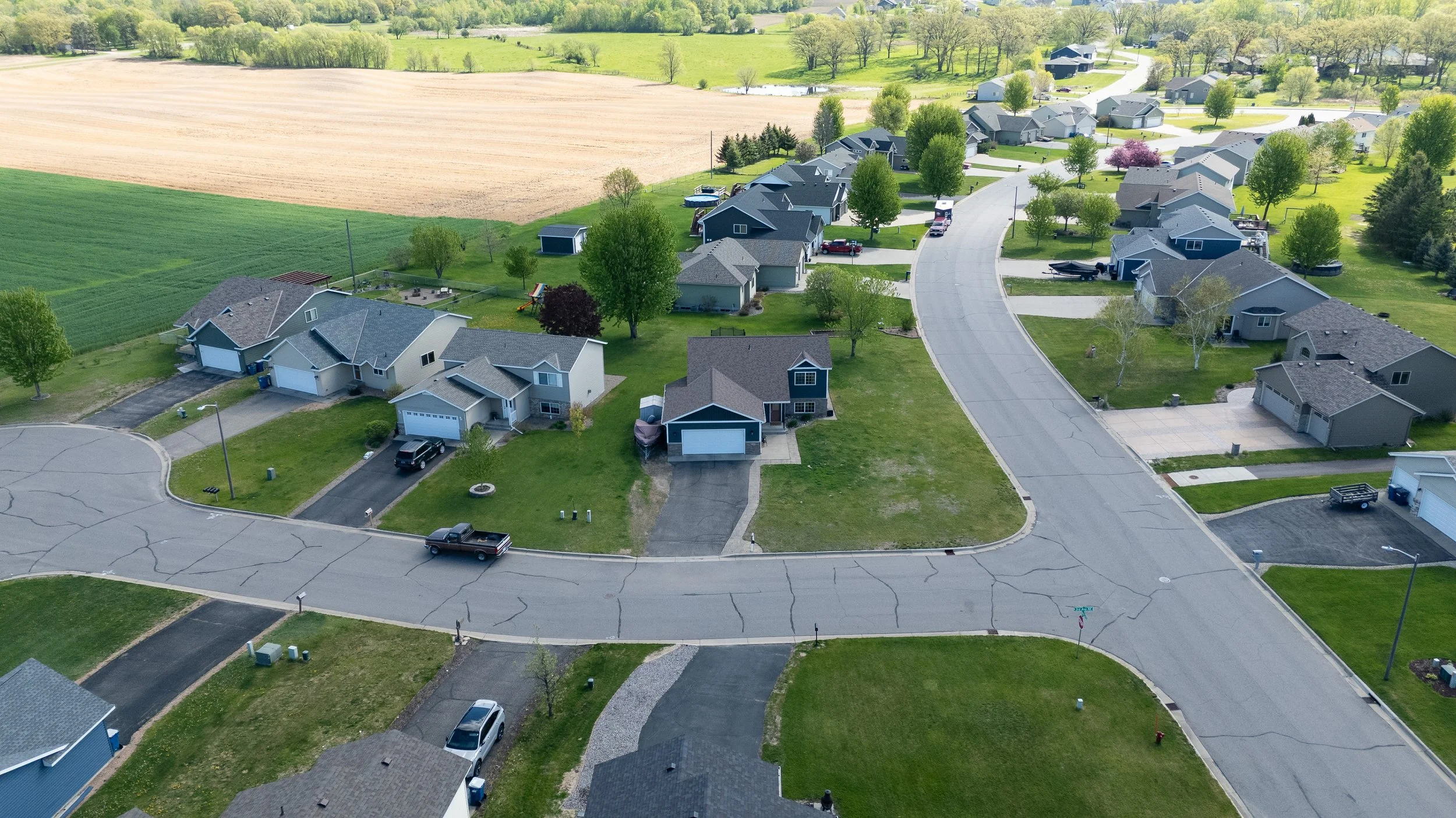 Aerial view of a suburban neighborhood with houses, trees, and a curved street, adjacent to open farmland with green fields and trees in the background.