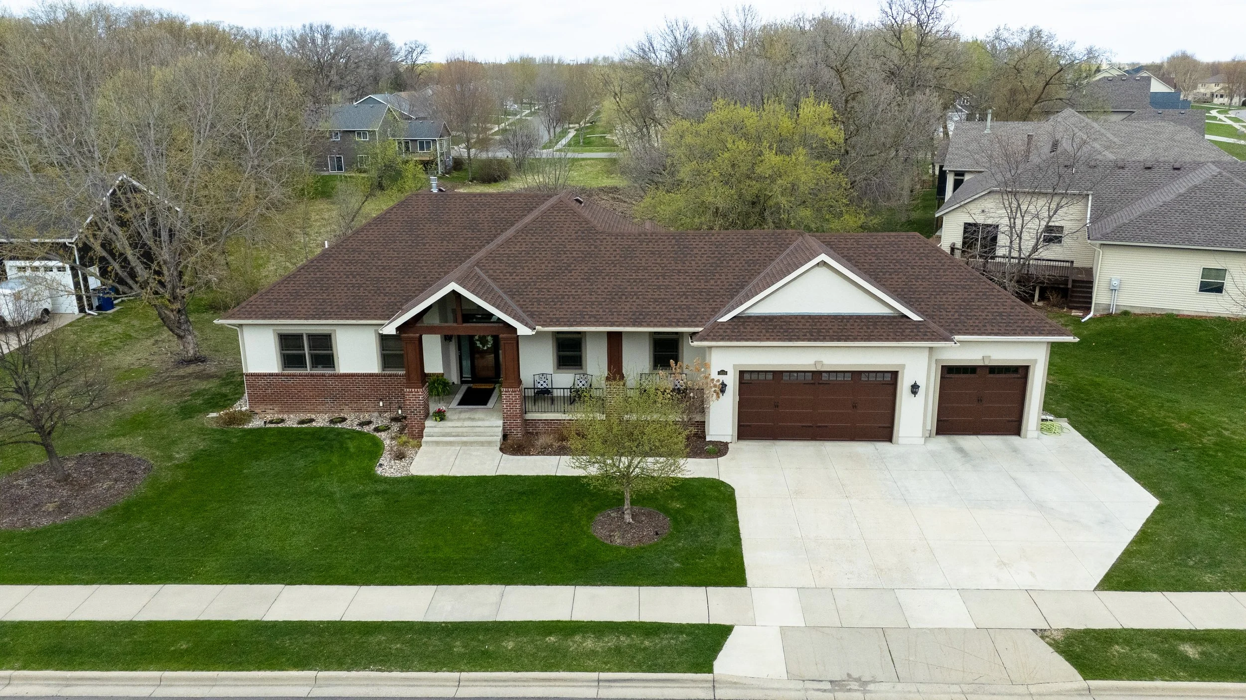 Aerial view of a suburban house with a brown roof, white walls, and a two-car garage, surrounded by a well-maintained lawn and trees, with neighboring houses and roads in the background.