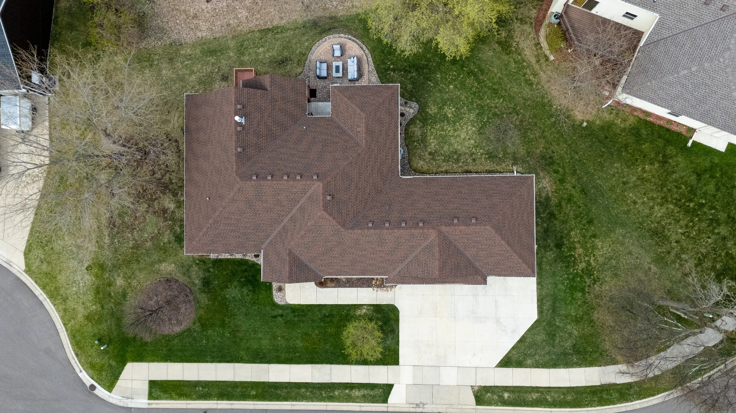 An aerial view of a house with a brown tiled roof, surrounding green yard, concrete pathway, and outdoor seating area with chairs and a table.
