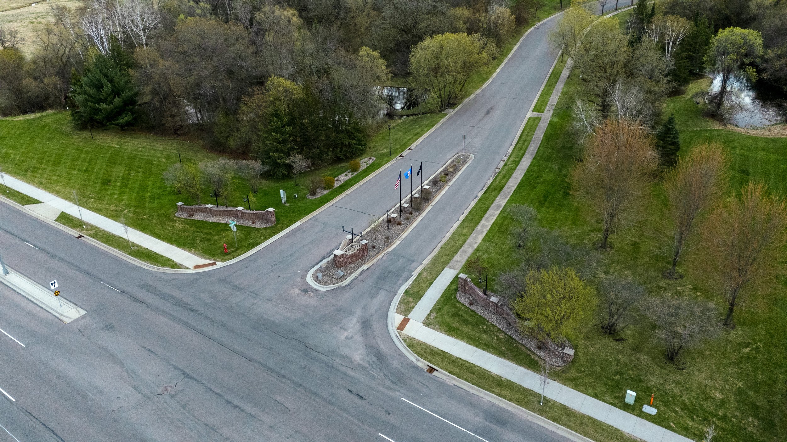 An aerial view of a park with a small road and sidewalk running through it, featuring flags, benches, parking meters, green lawns, trees, and a small water body surrounded by trees.