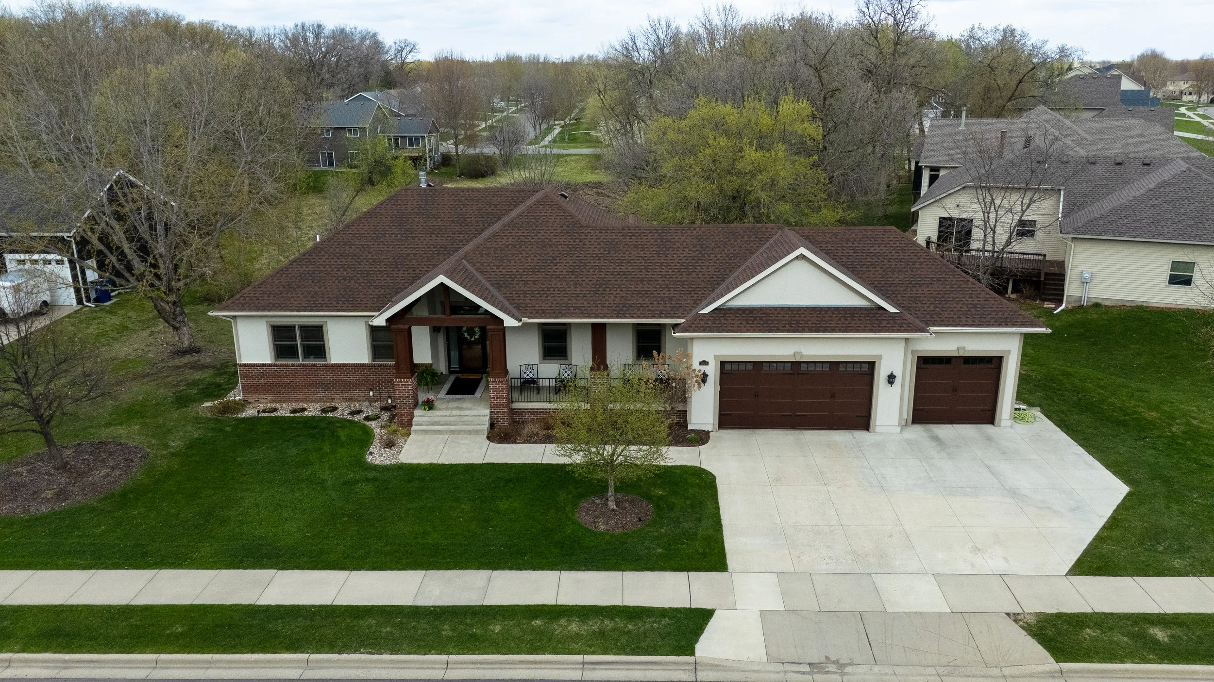 A suburban house with a brick and white exterior, brown roof, attached two-car garage, front porch with a small set of stairs, and manicured lawn with young trees. Surrounding houses are visible in the background among leafless trees, indicating earl