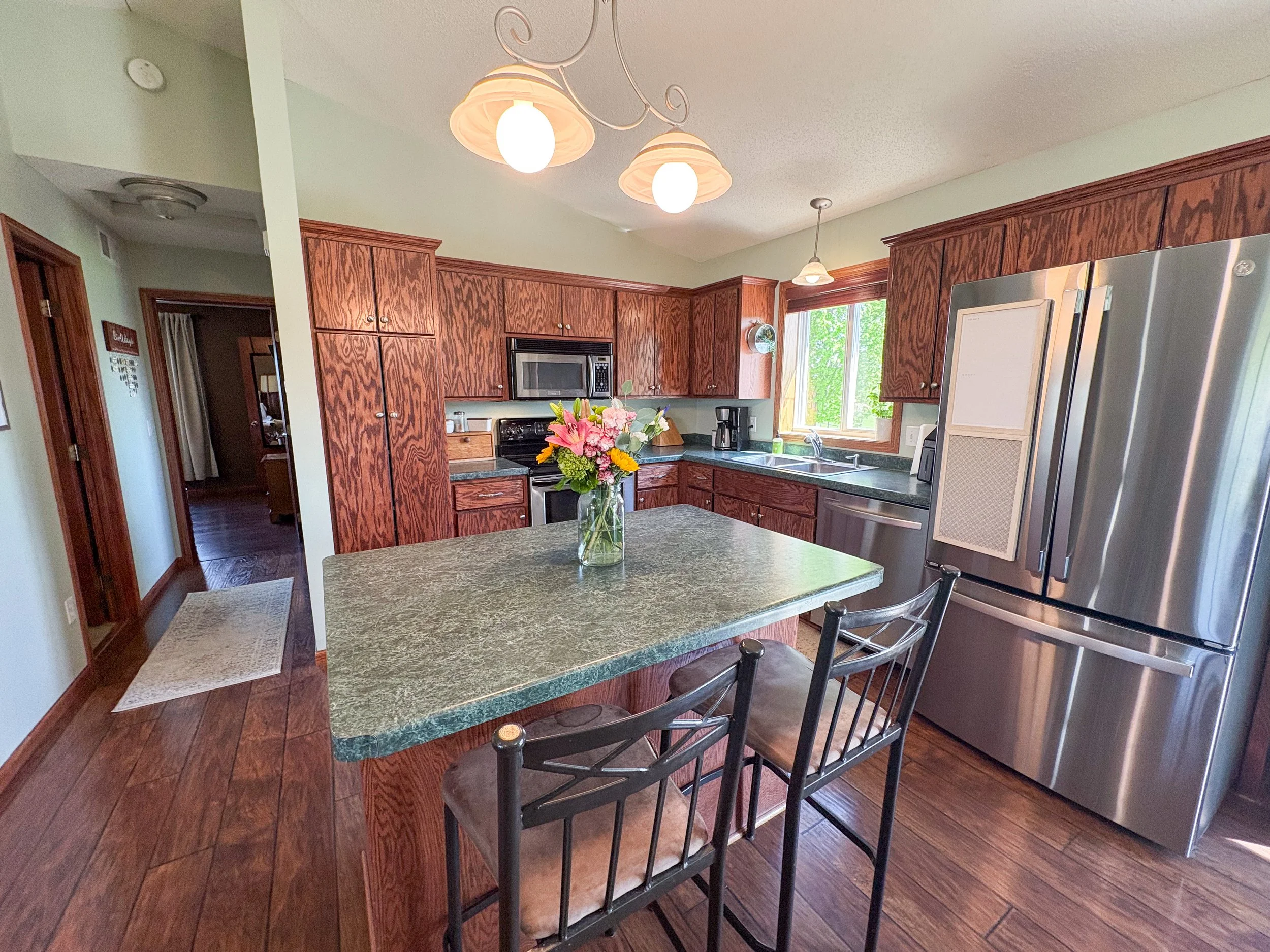 Kitchen with wooden cabinets, a kitchen island with a vase of flowers, a stainless steel refrigerator, a microwave, and a window with greenery outside.