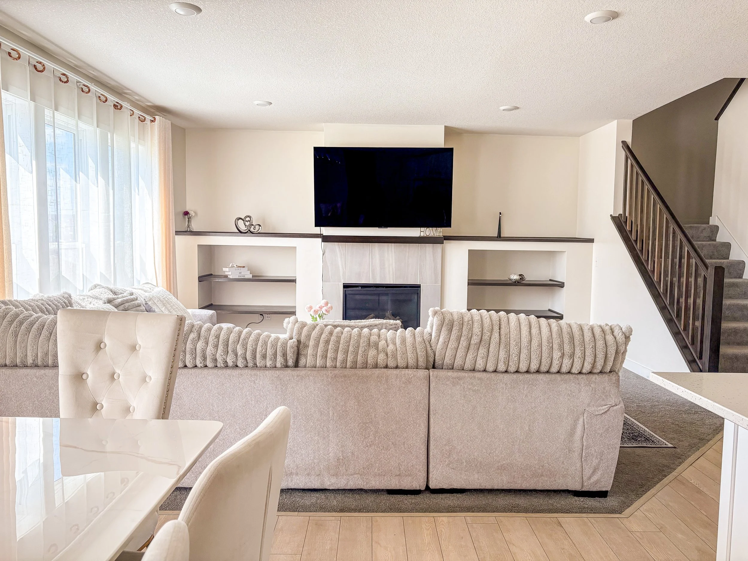 A spacious living room with a beige sofa, a white dining table with chairs, a built-in fireplace with shelves, a large flat-screen TV above the fireplace, and a staircase to the right. There's a window with sheer curtains on the left side.