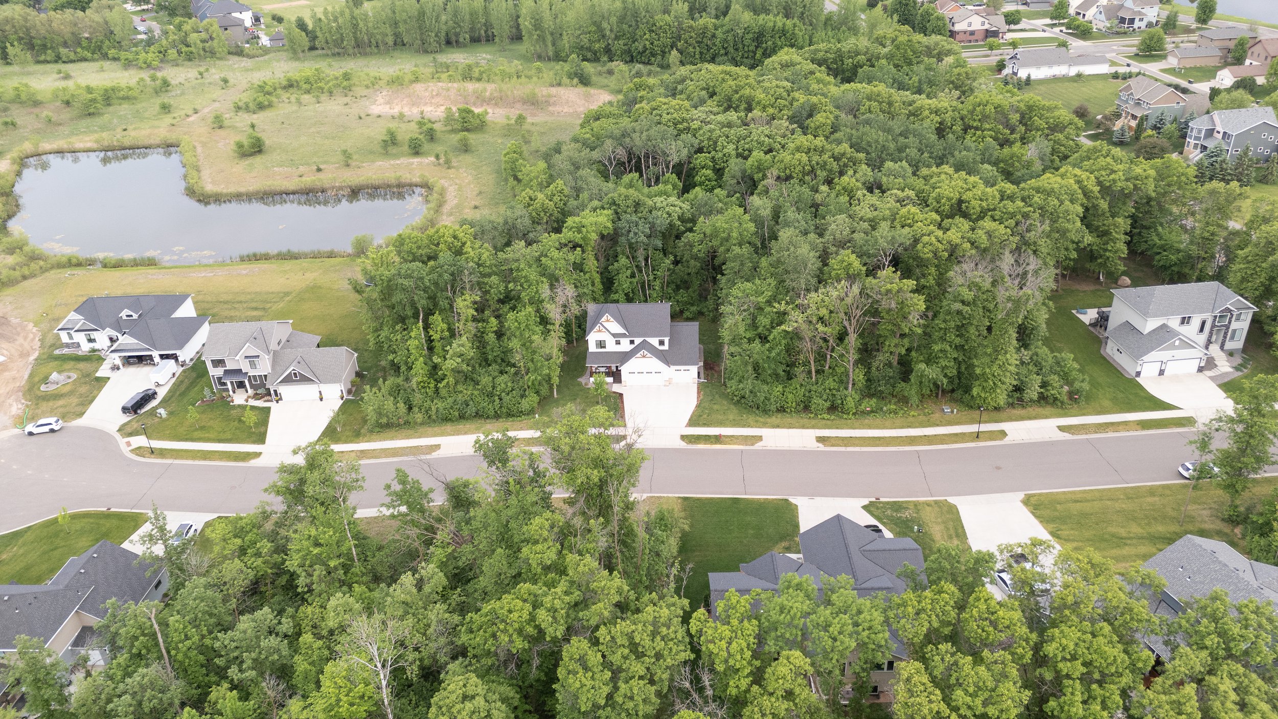 Aerial view of a suburban neighborhood with several houses, trees, and a pond, surrounded by greenery.