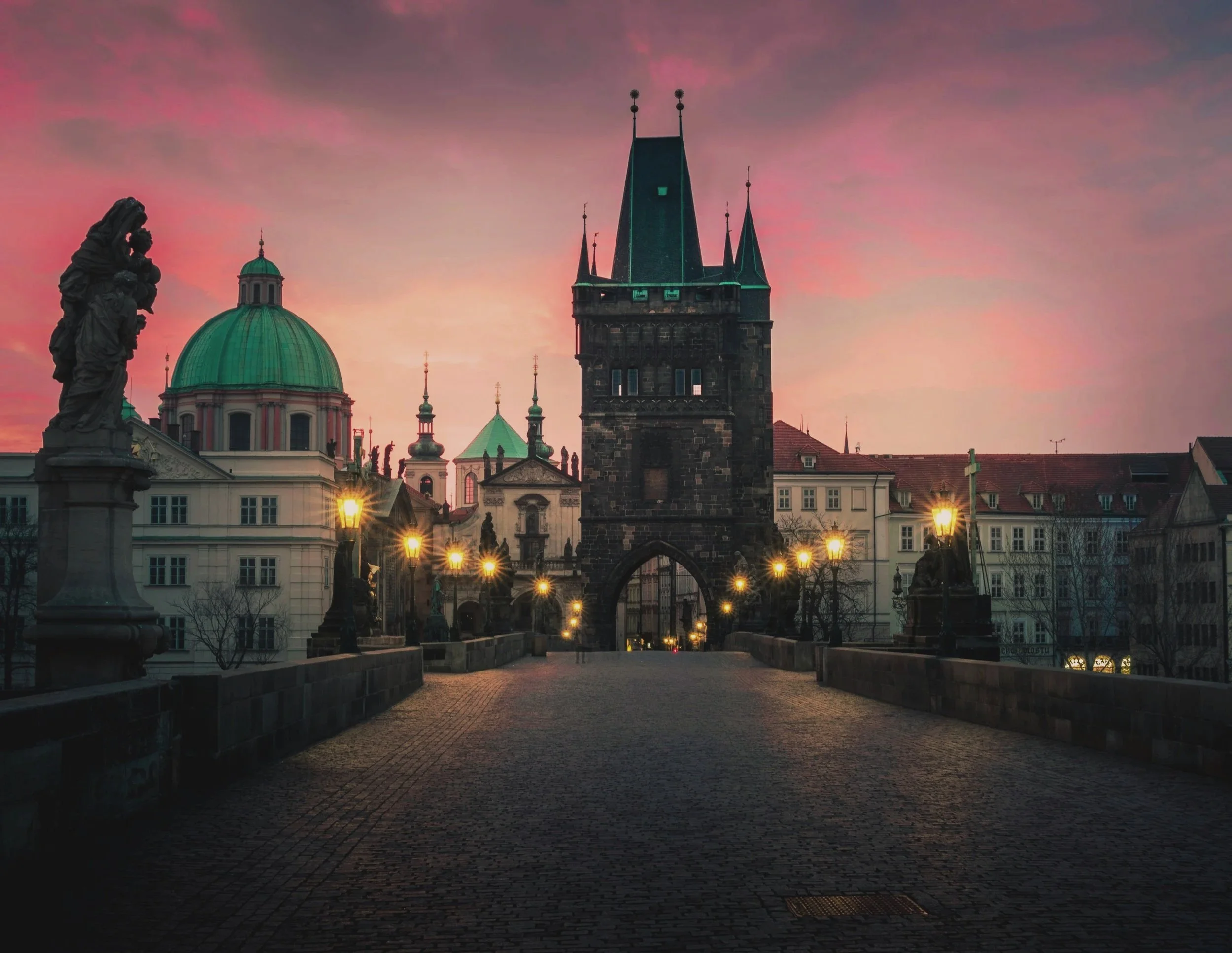 Charles Bridge, Prague - at dusk with a pink and purple sky, featuring historic European buildings, gothic spires, street lamps illuminating a cobblestone pathway.