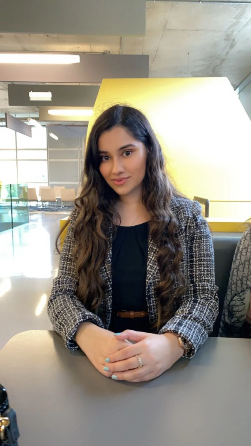 A young woman with long, curly brown hair sitting at a table in a modern, well-lit space, wearing a patterned blazer and a black top.