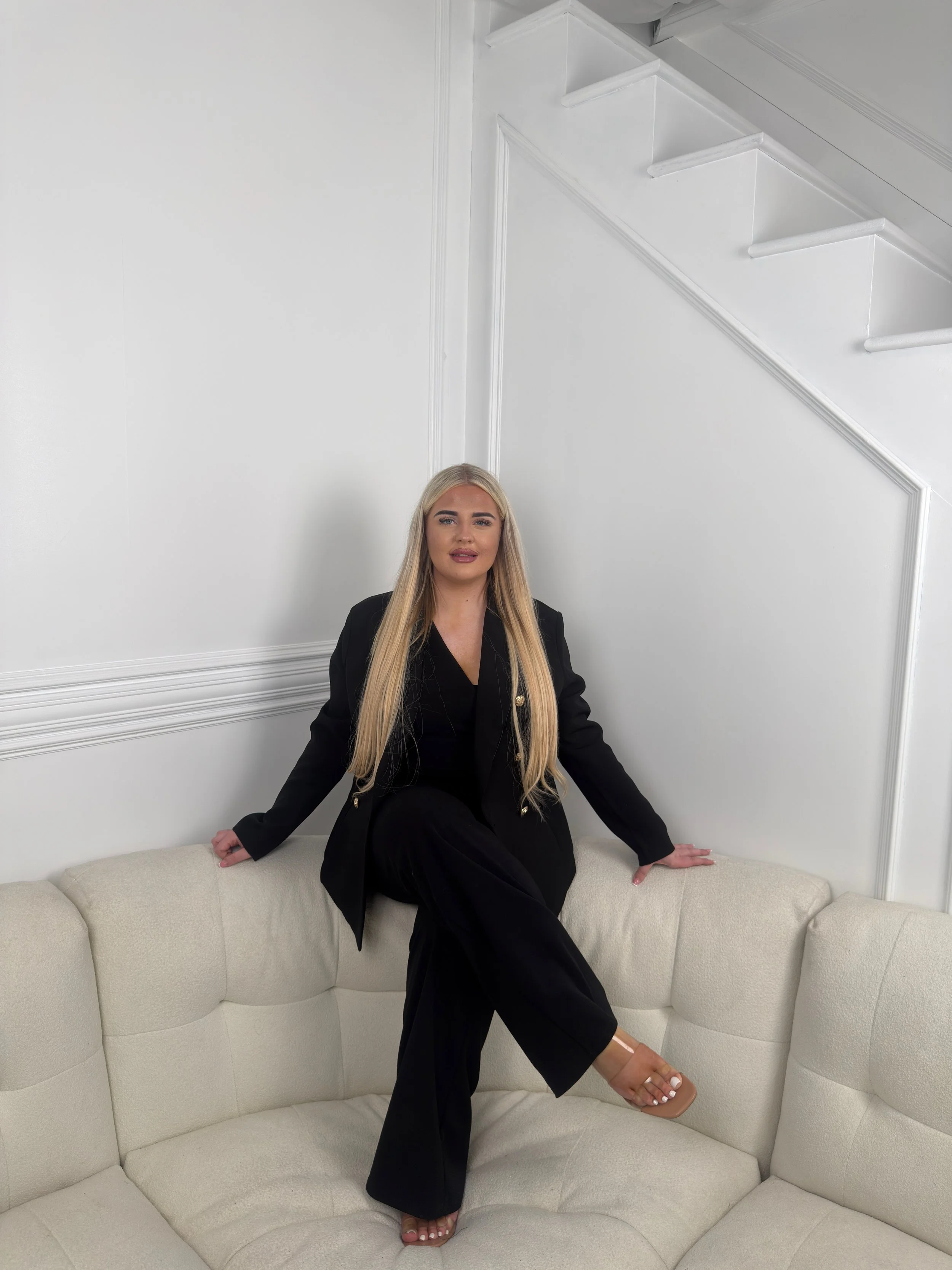 A woman with long blonde hair sitting on a cream-colored curved sofa, wearing a black blazer and pants, with one leg crossed over the other. She is positioned in front of a white wall with decorative paneling and a staircase visible at the top.