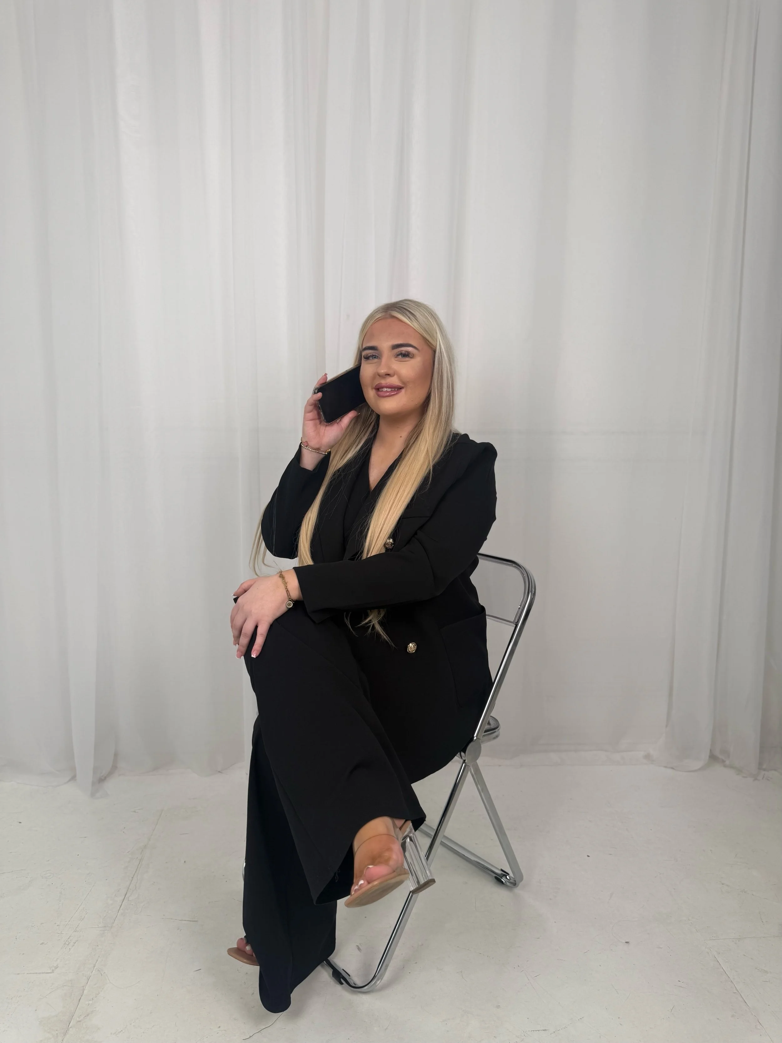 A woman with long blonde hair, dressed in a black blazer and black pants, sitting on a clear plastic chair in front of a white curtain. She is holding a phone to her ear with her right hand and is smiling.