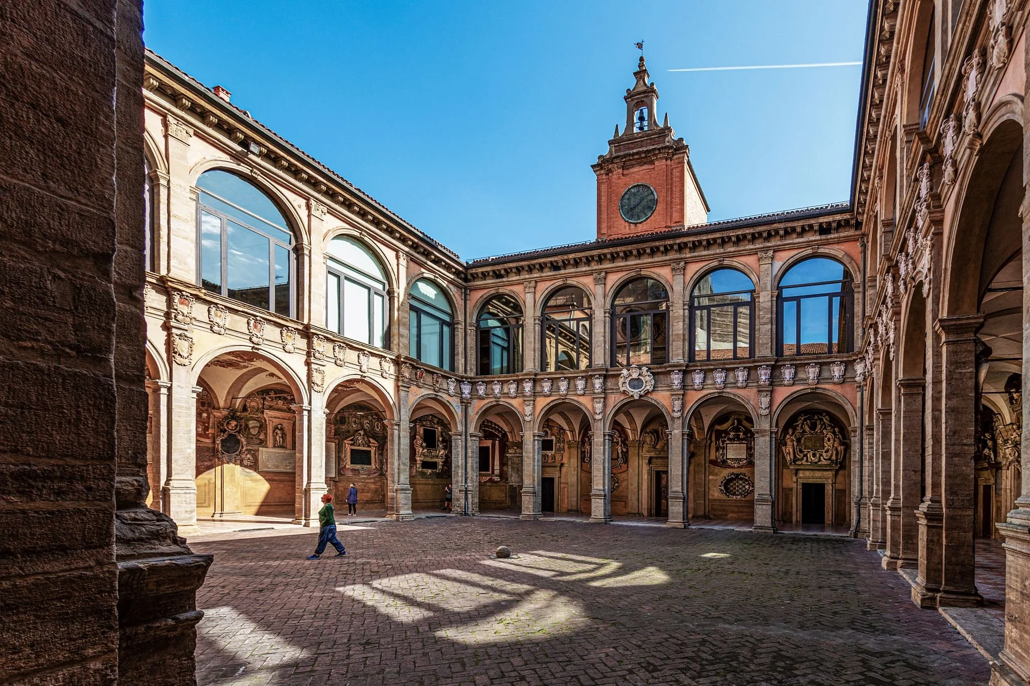 Pátio interno de um prédio antigo com arcos, colunas e uma torre com relógio ao centro. Há algumas pessoas caminhando no chão de pedra.