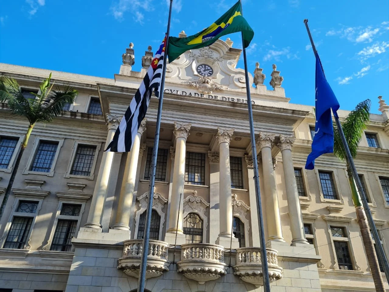 Prédio antigo de fachada clássica com colunas dóricas, com três bandeiras hasteadas na frente. Uma delas é a bandeira de São Paulo, com listras pretas e brancas e uma estrela vermelha, ao centro. A fachada tem detalhes decorativos entre as janelas e um relógio no topo. Céu azul ao fundo.