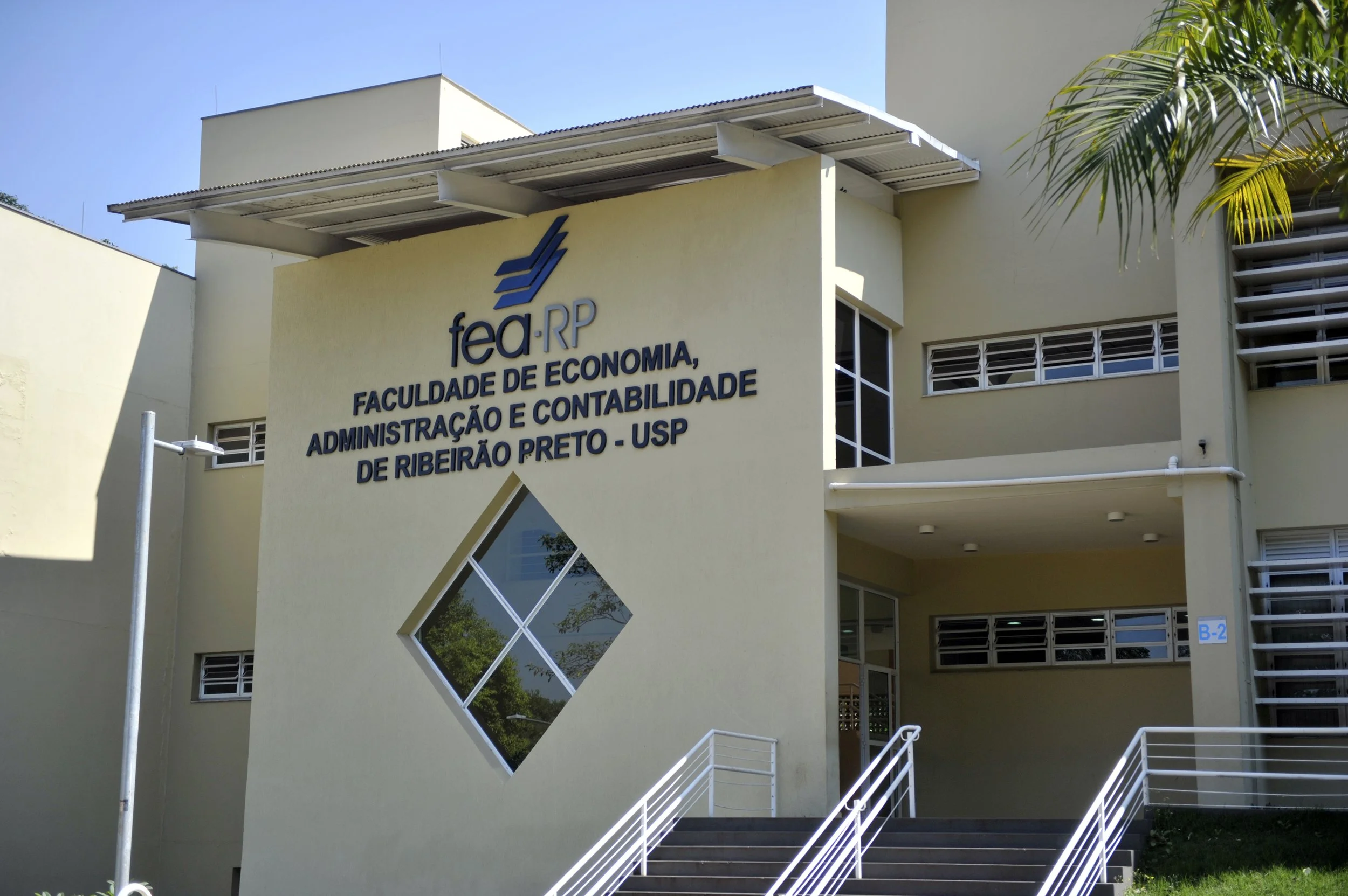 Fachada do prédio da Faculdade de Economia, Administração e Contabilidade de Ribeirão Preto da USP, com paredes amarelas, janelas com venezianas, escadas e viseiras no teto, parte de uma árvore ao lado e céu claro ao fundo.