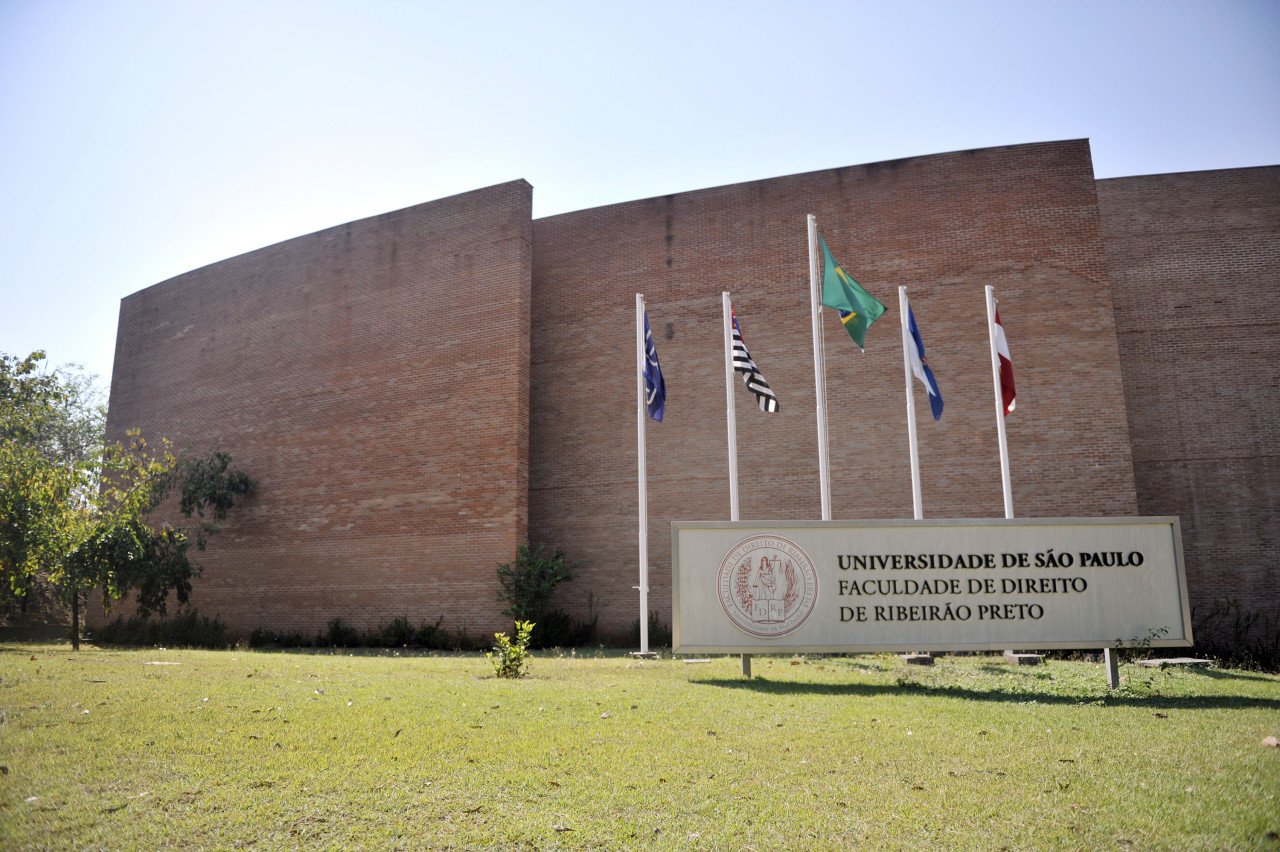 Prédio de tijolos com cinco bandeiras em postes na frente e placa de sinalização indicando a Faculdade de Direito da Universidade de São Paulo de Ribeirão Preto.