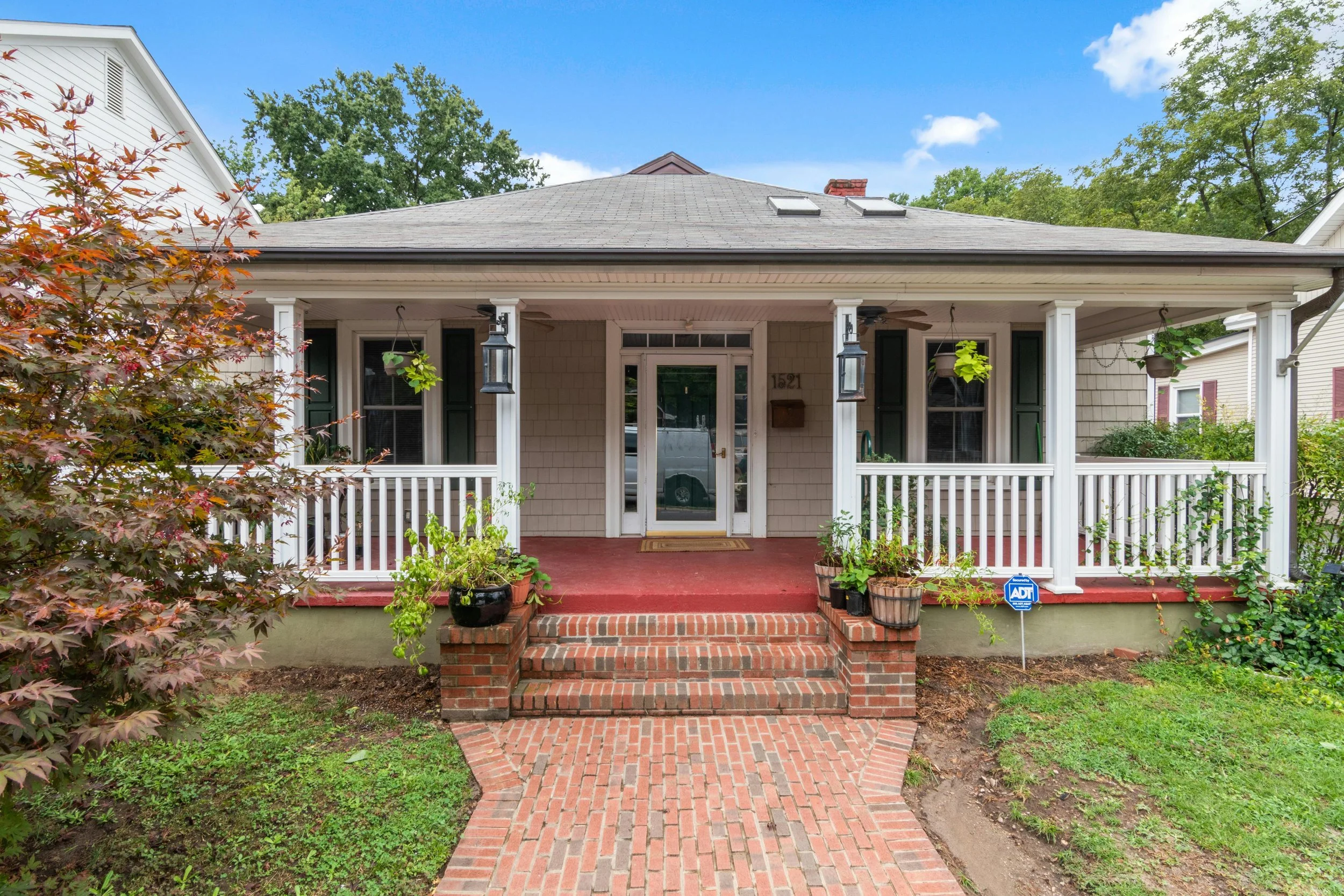 Front porch of a house with brick steps leading up to a white door, surrounded by a white railing, with hanging plants and potted plants, and a red brick floor. There is a tree with red leaves on the left and green trees in the background, with a bright blue sky.