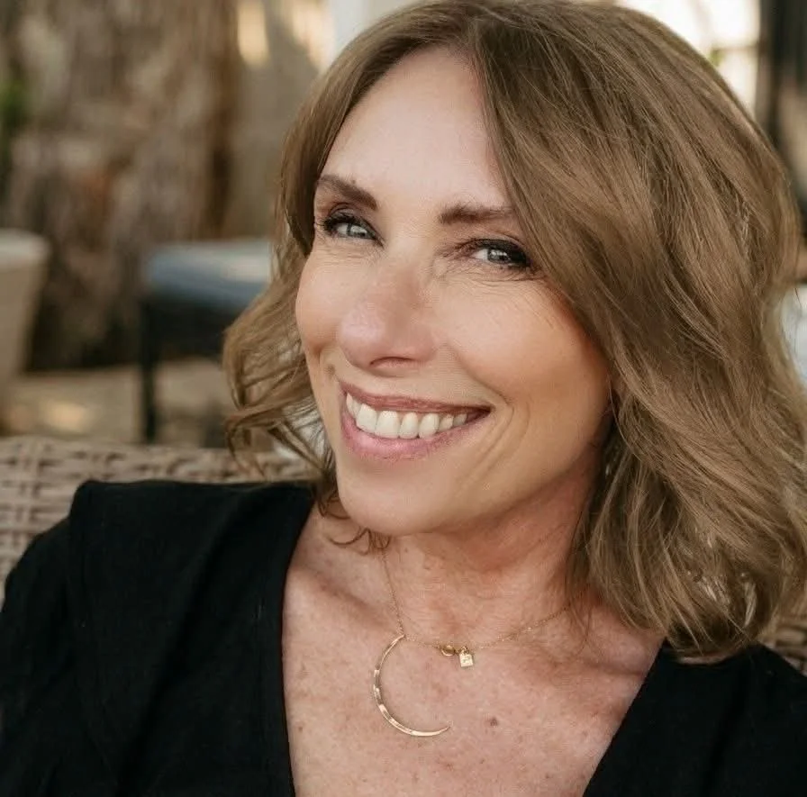 A woman with shoulder-length wavy light brown hair, smiling softly, wearing a black top and a gold necklace with a crescent moon pendant, sitting outdoors on a wicker chair.