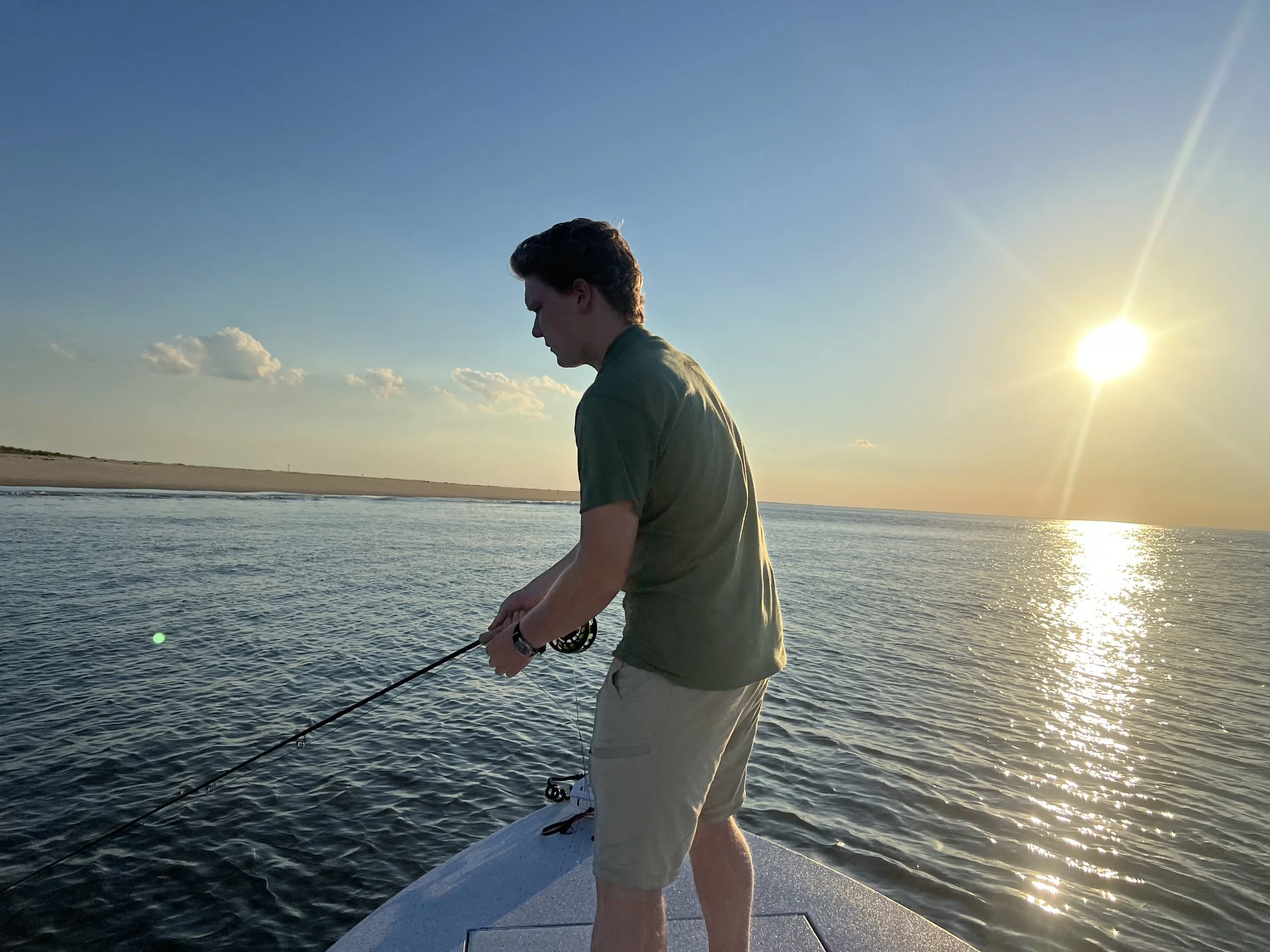 A person standing on a boat, fishing in the water during sunset, with the sun low in the sky and clouds in the background.
