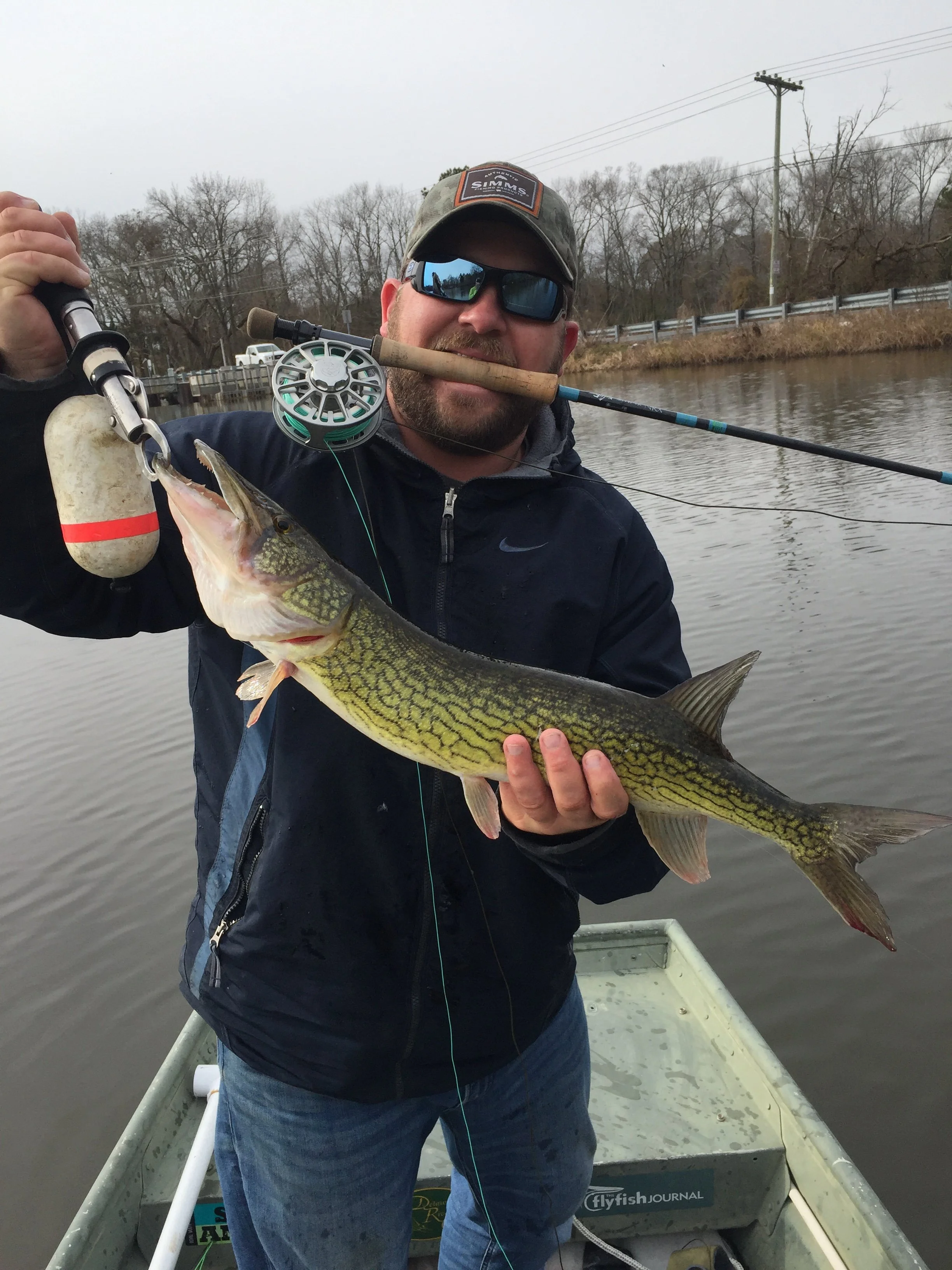 Man in sunglasses and a baseball cap holding a largemouth bass in a boat on a lake with bare trees and power lines in the background.