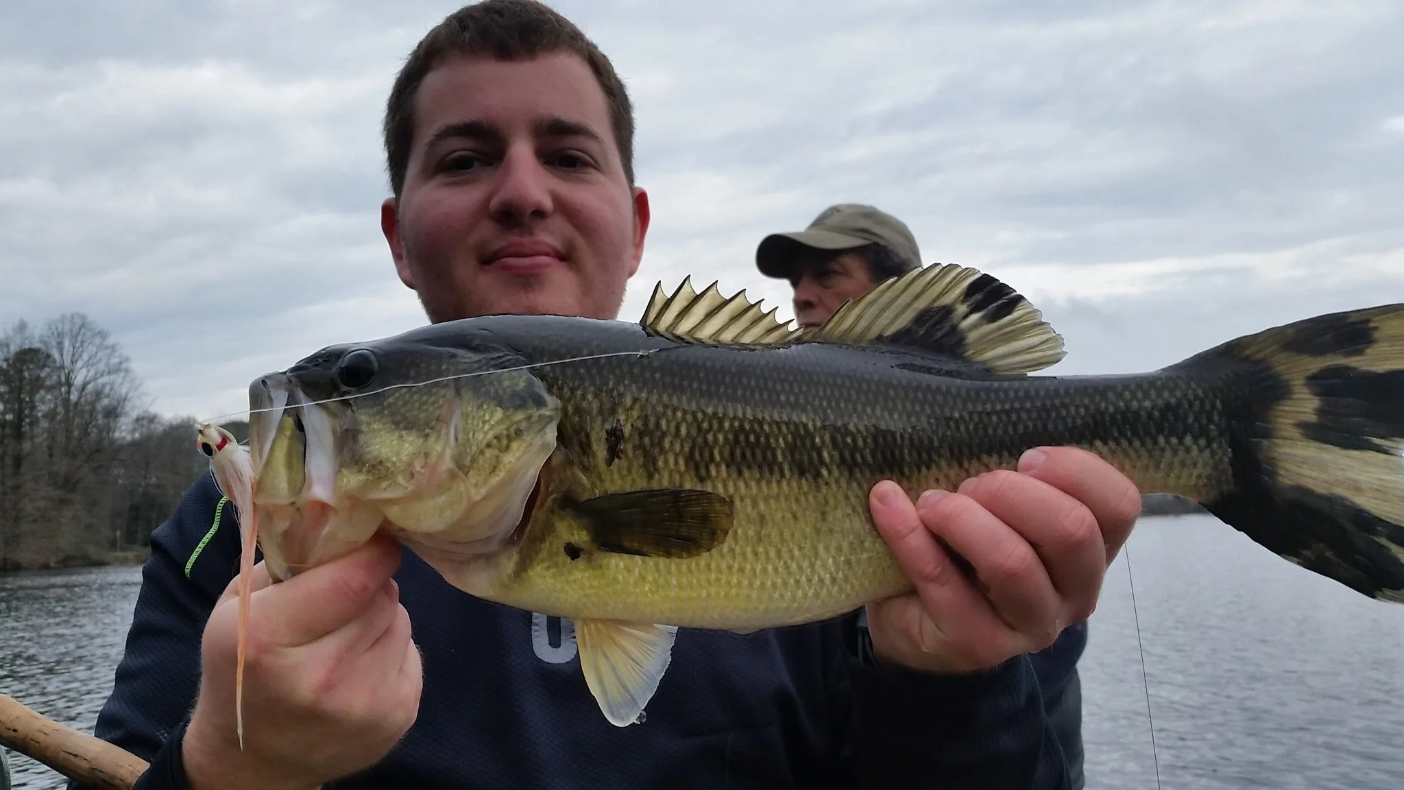 A young man holds a large fish with dark scales and a yellowish belly, posing outdoors near water, with another man in the background.