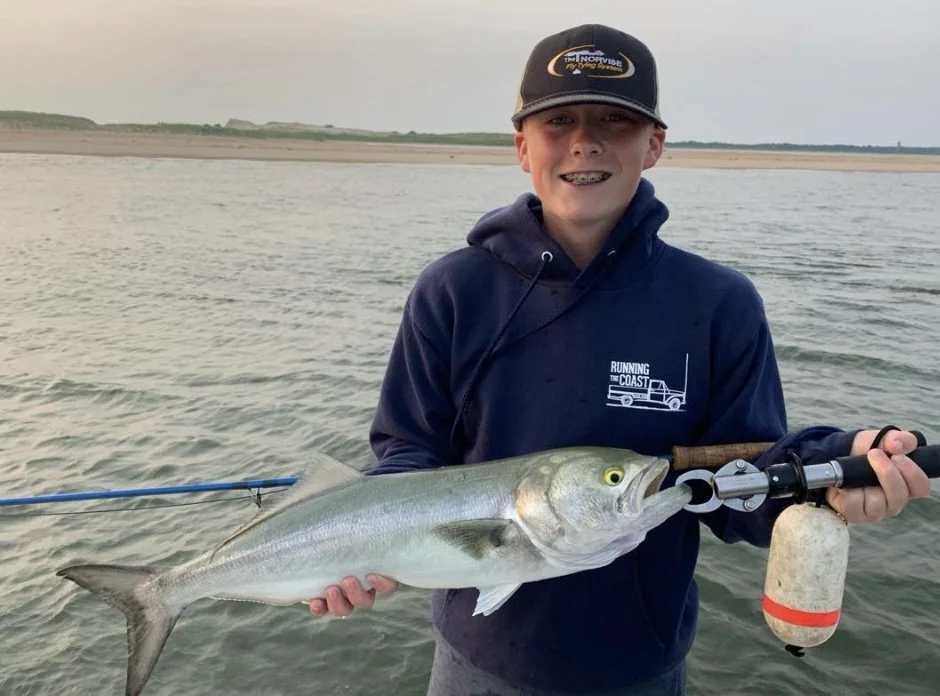 Young boy holding a fish with a fishing line and tool, standing in a body of water on a cloudy day, wearing a navy hoodie and black cap.