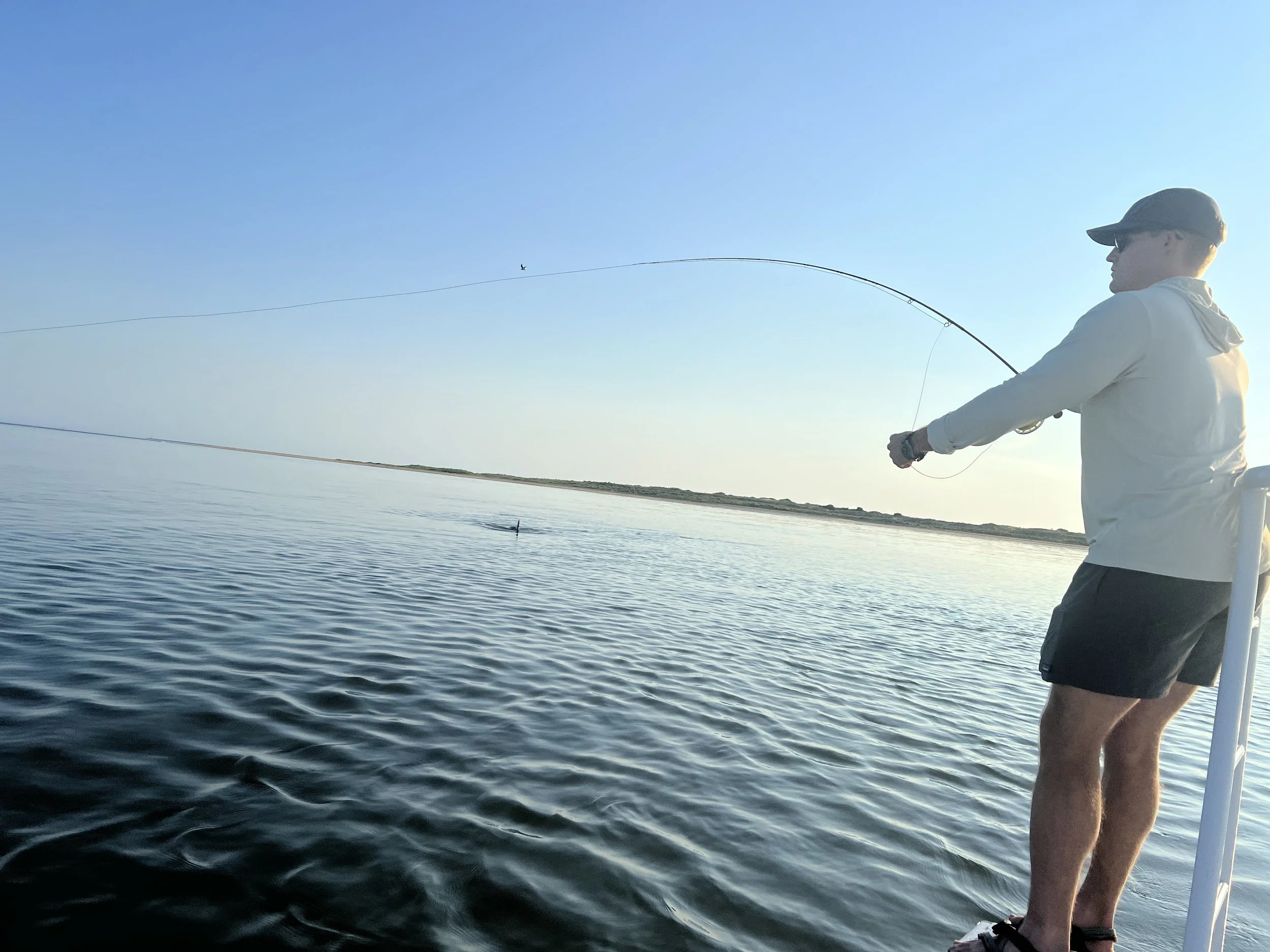 A person in a white hoodie, sunglasses, and shorts stands on a boat fishing in a body of water during the daytime. The person is holding a fishing rod, and a small fish is seen jumping out of the water.