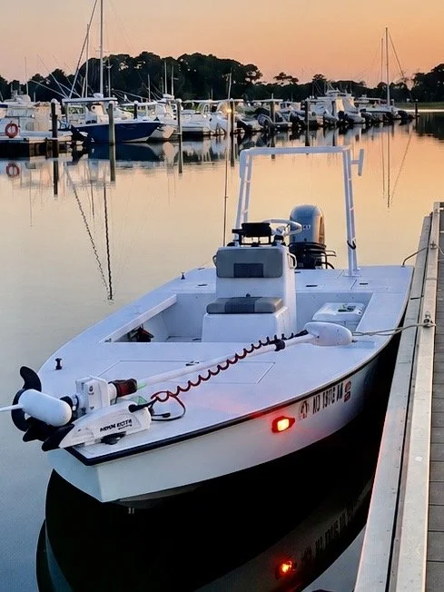 A white boat docked at a marina during sunset, with other boats in the background and calm water reflecting the scene.