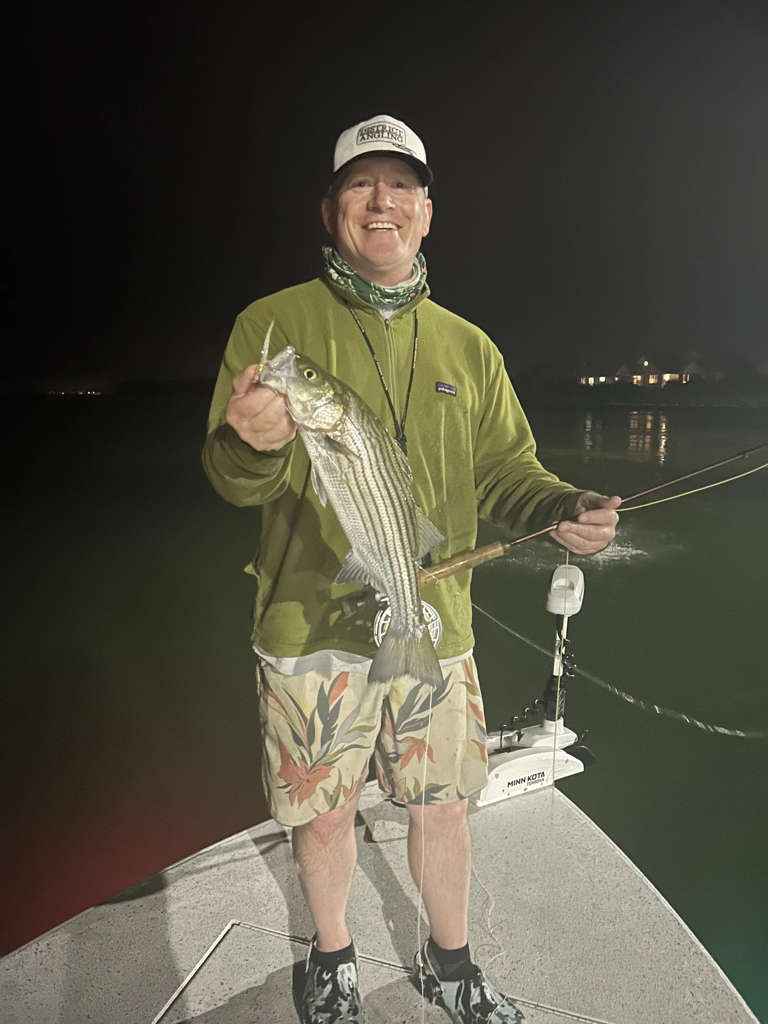 A man standing on a dock at night holding a large fish in one hand and a fishing rod in the other. He is wearing a hat, a green jacket, floral shorts, and boots, smiling at the camera with city lights visible in the background.