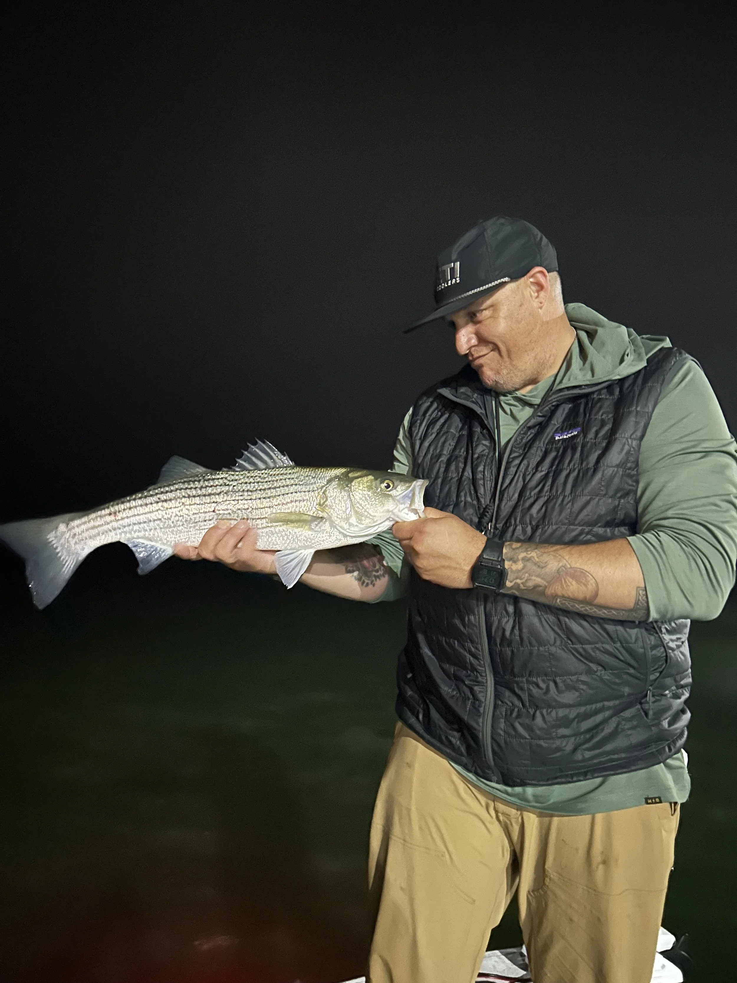A man wearing a black cap, green hoodie, black vest, and khaki pants, holding a large fish at night.