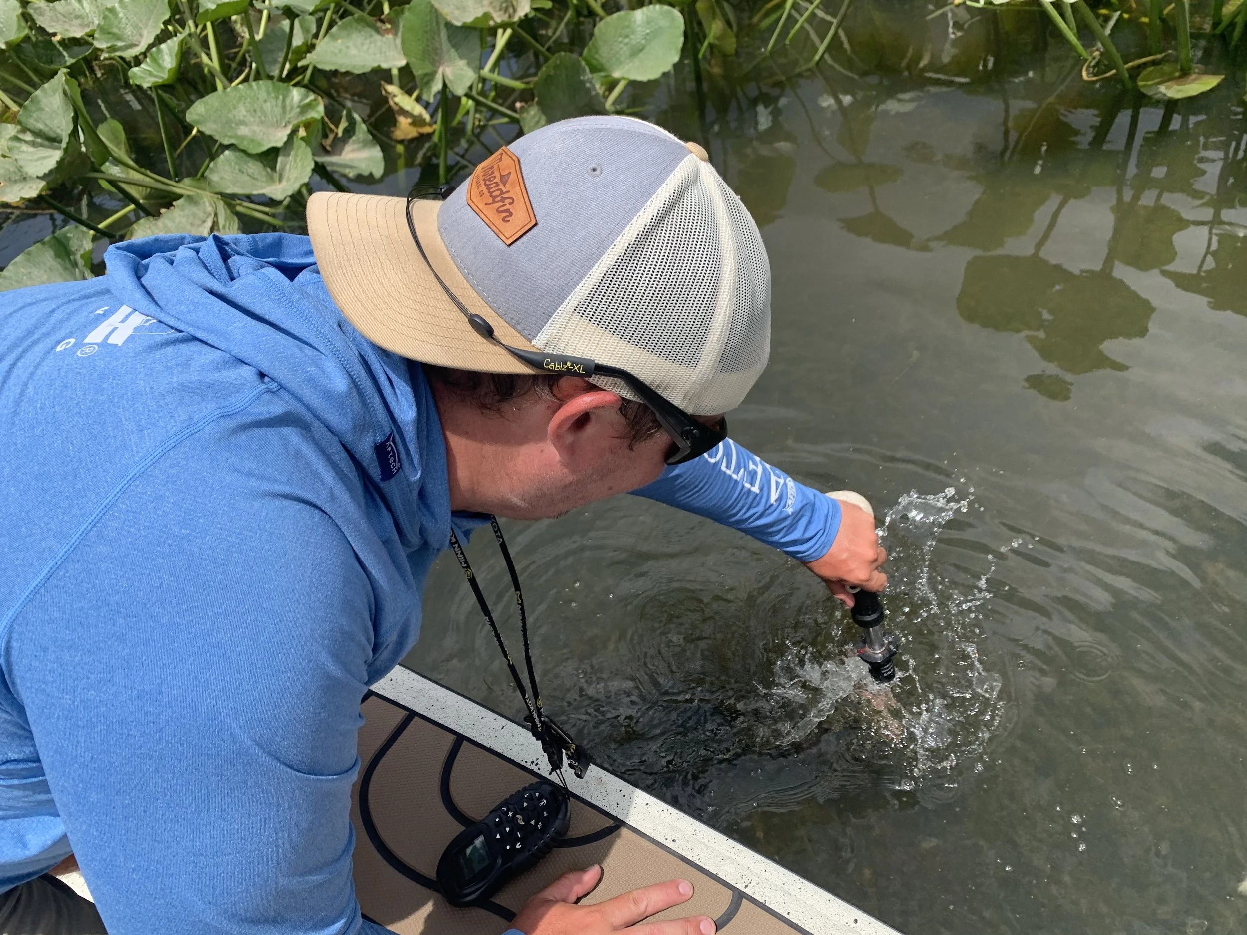 A person in a blue hoodie and a cap with a mesh back is standing on a boat, casting a fishing rod into a body of water surrounded by green lily pads.