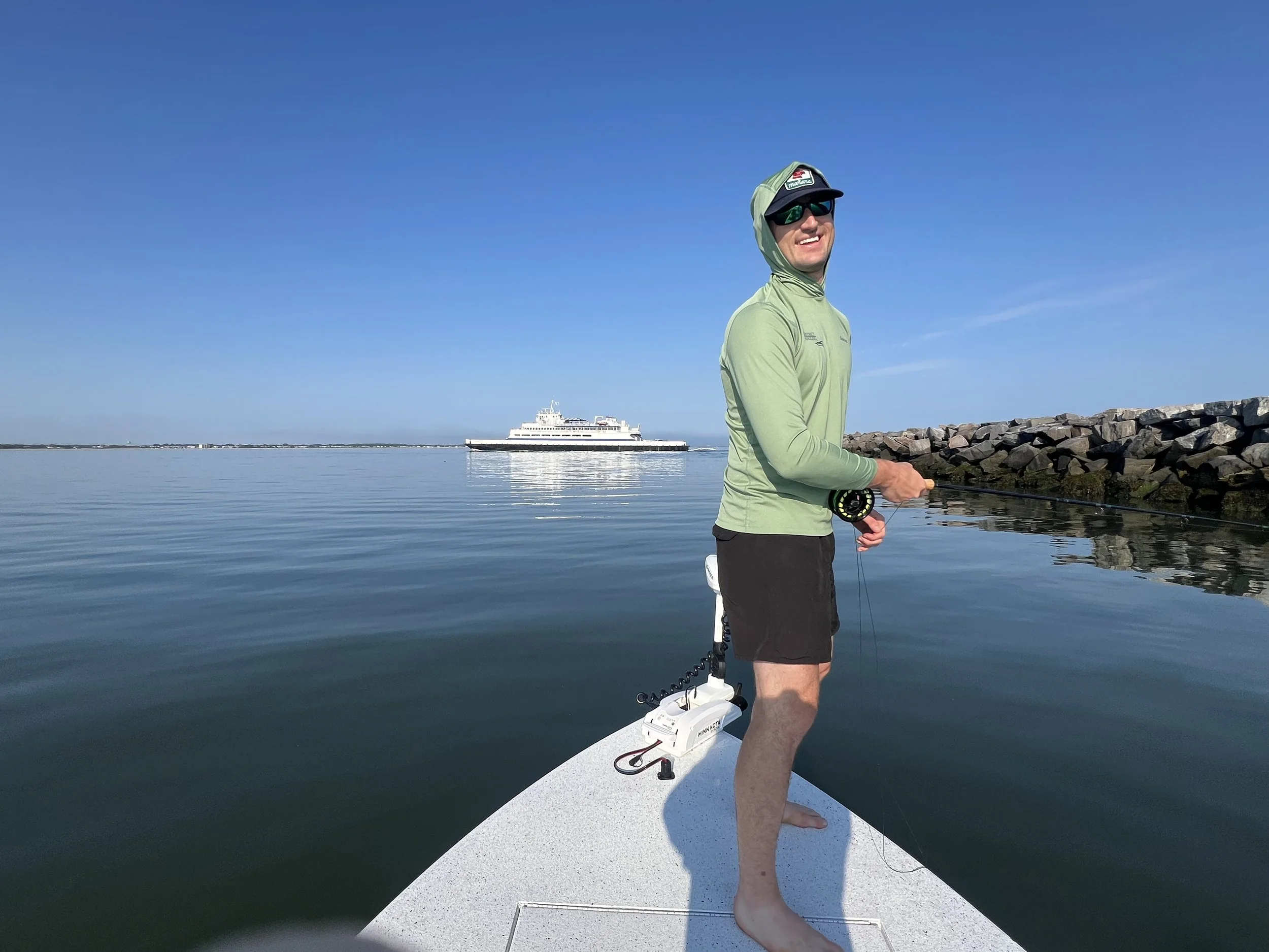 Man standing on a paddleboard on calm water, smiling, wearing a green hoodie, black shorts, sunglasses, and a cap with a fishing rod in hand. In the background, a large white ferry boat and a rocky shoreline are visible under a clear blue sky.