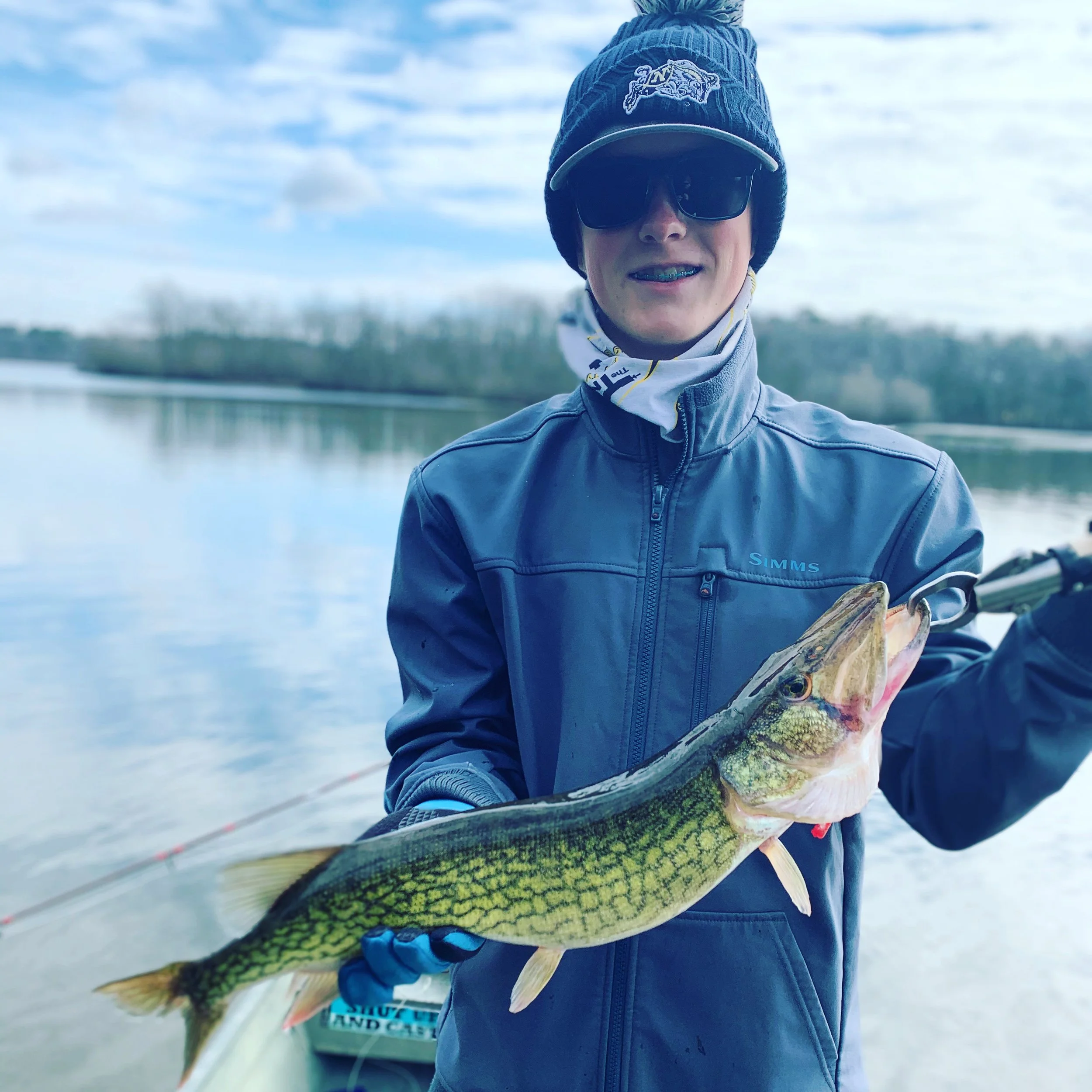 A person wearing a navy blue jacket, black sunglasses, and a blue beanie hat is holding a fish with green and yellow scales near a body of water. The person is outdoors on a cloudy day.