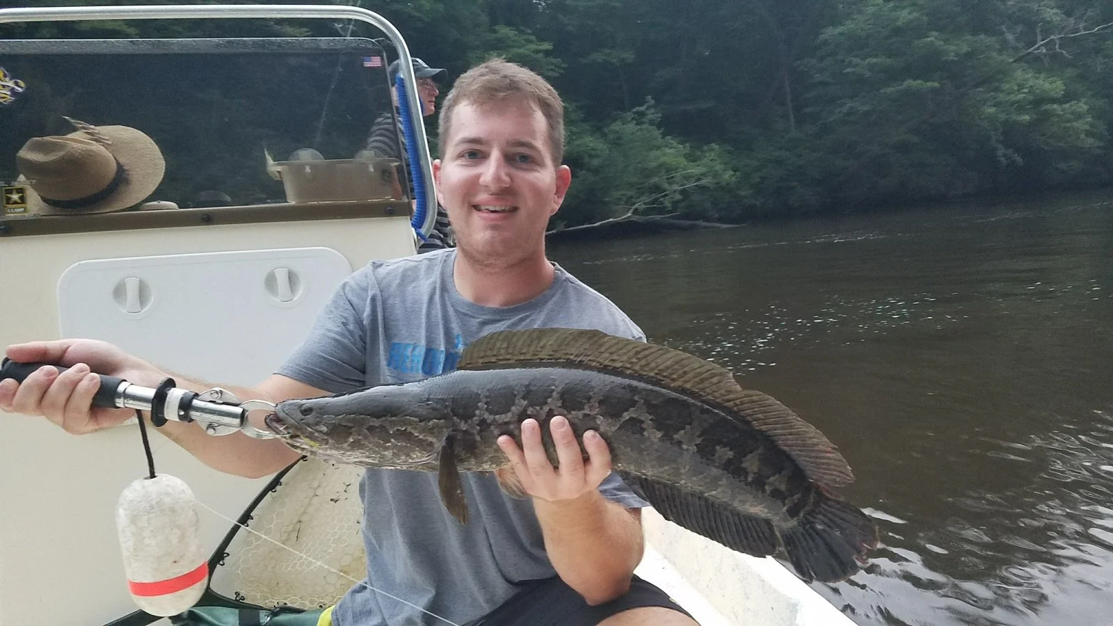 Young man holding a large fish on a boat by a river, with green trees in the background.