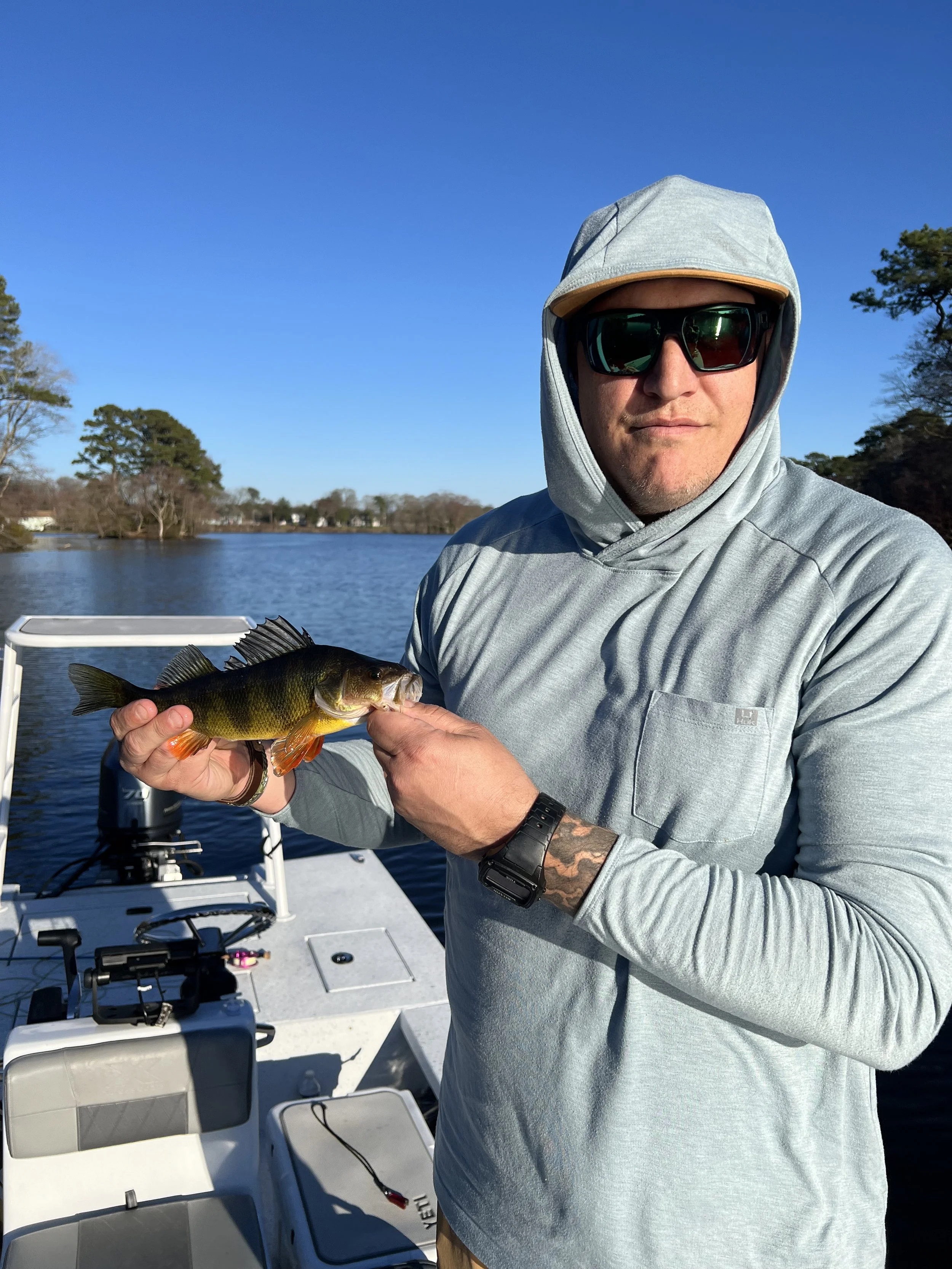Man wearing sunglasses and a gray hoodie, holding a large fish on a boat in a lake with trees and blue sky in the background.