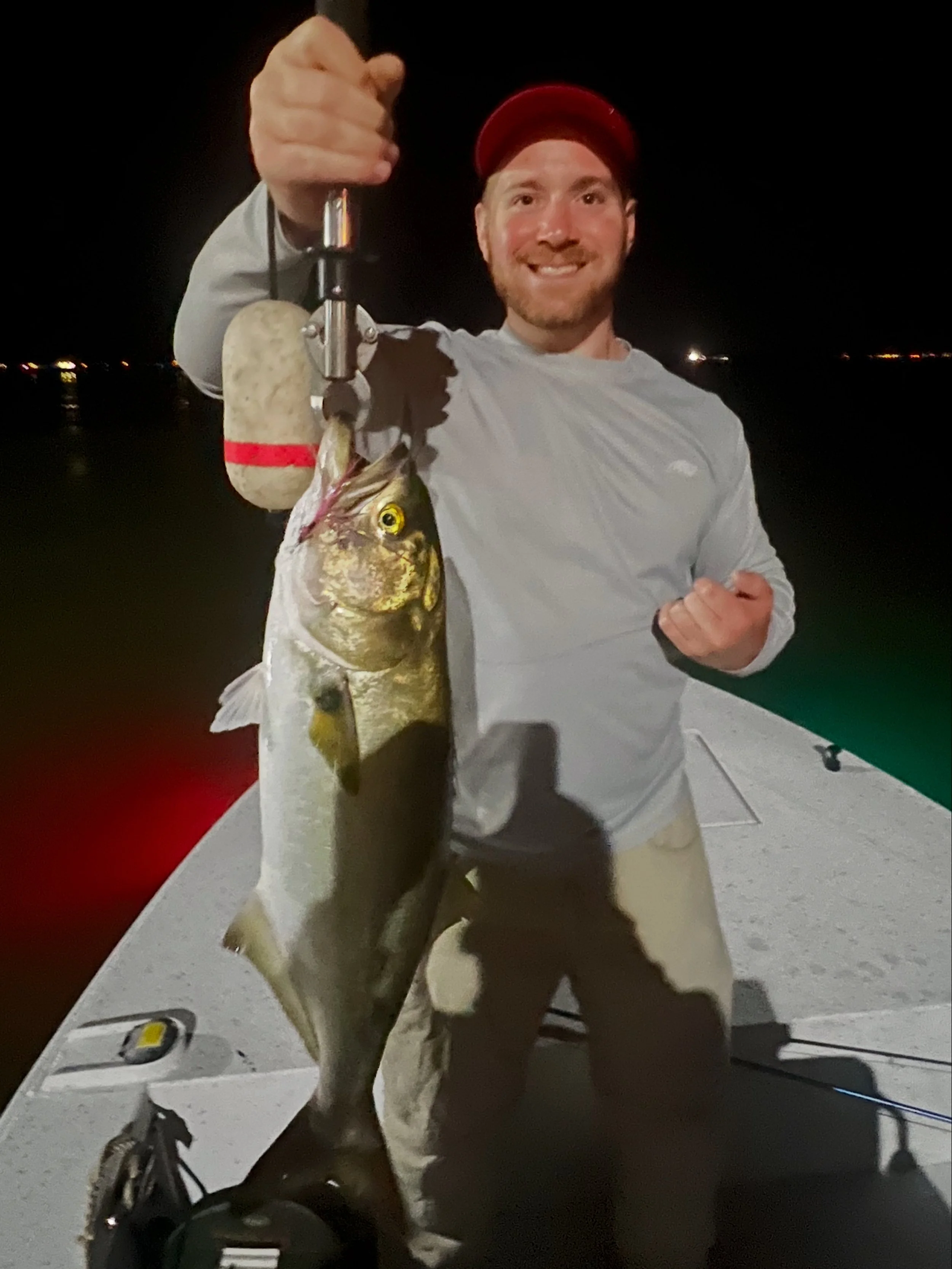 A man wearing a red cap and gray long-sleeve shirt smiling and holding a fish by a fishing line at night with water and city lights in the background.