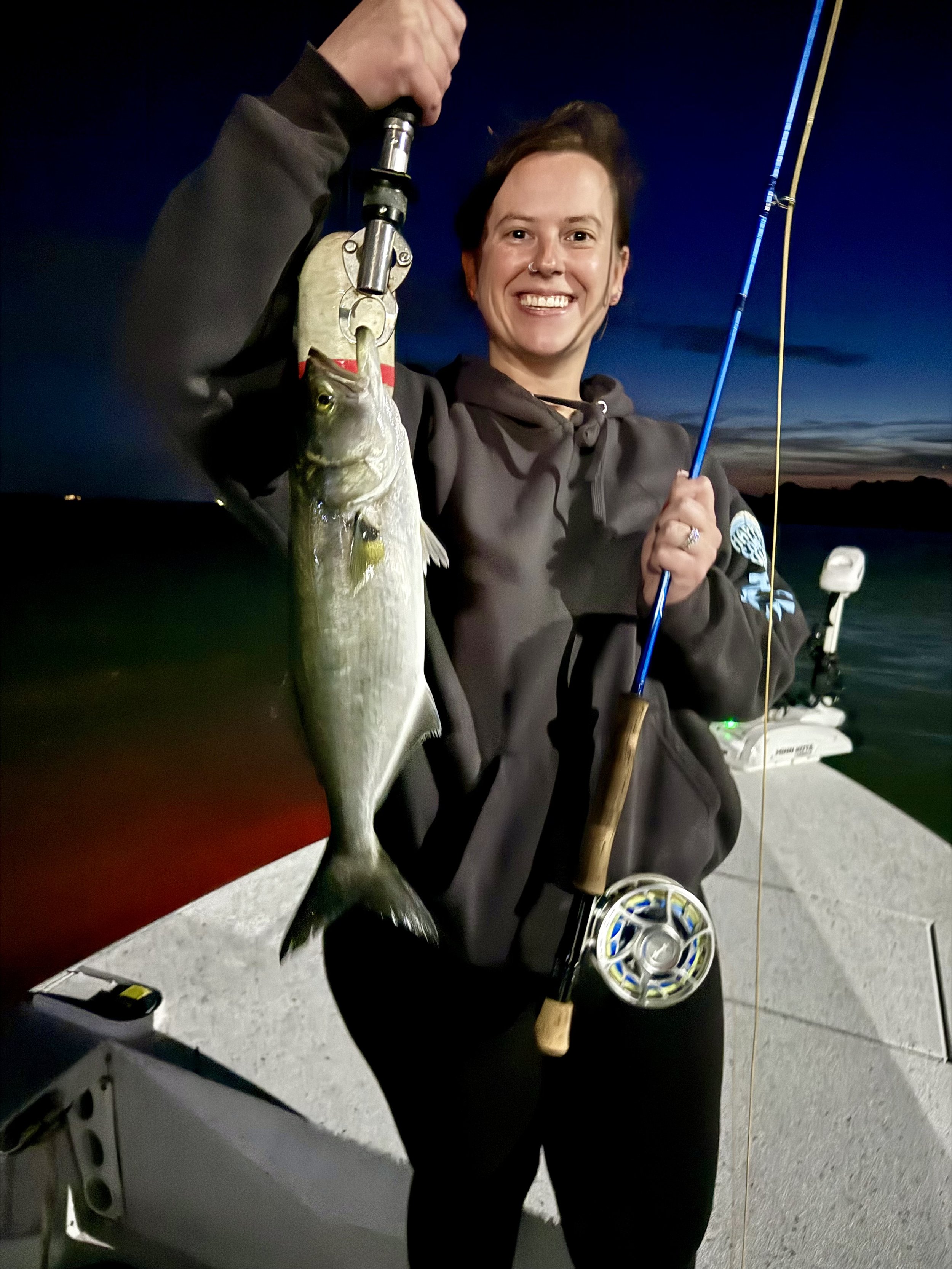 Person smiling on a boat at dusk holding a fishing rod and a freshly caught fish.