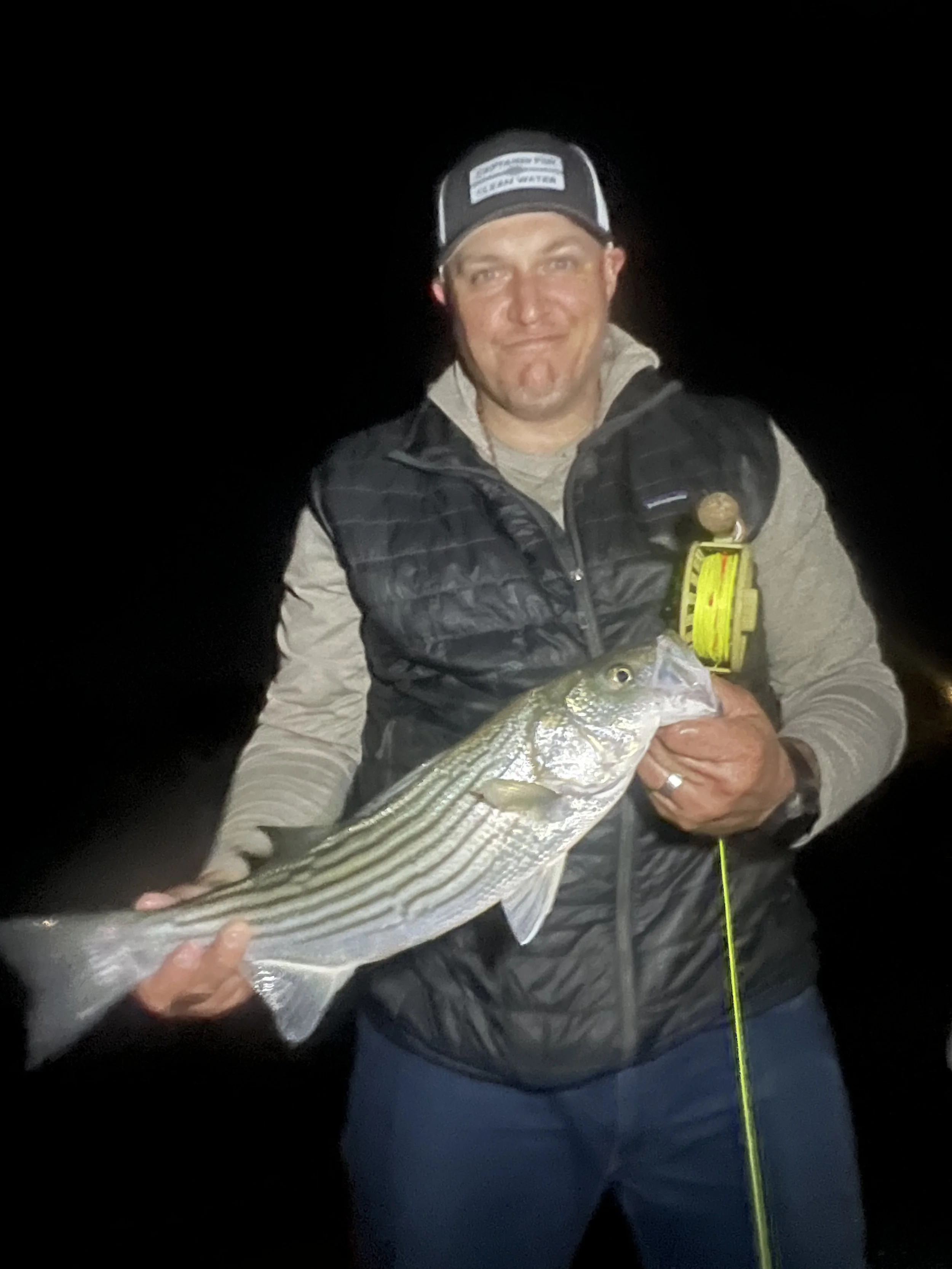 Man in dark jacket and baseball cap holding a large striped fish at night