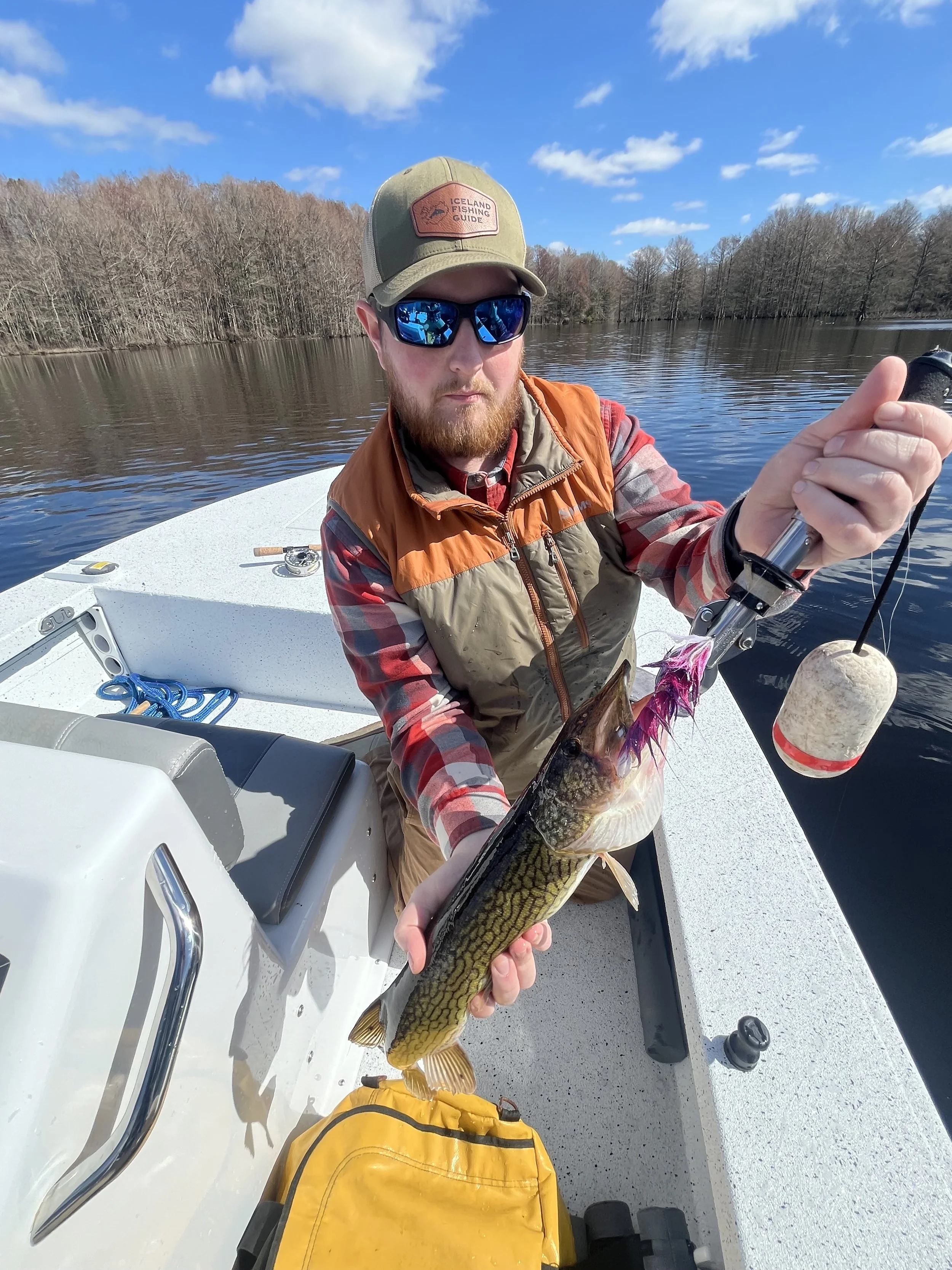 Man in a boat holding a fish he caught, wearing sunglasses, a cap, and a vest, with a background of water and trees under a partly cloudy sky.