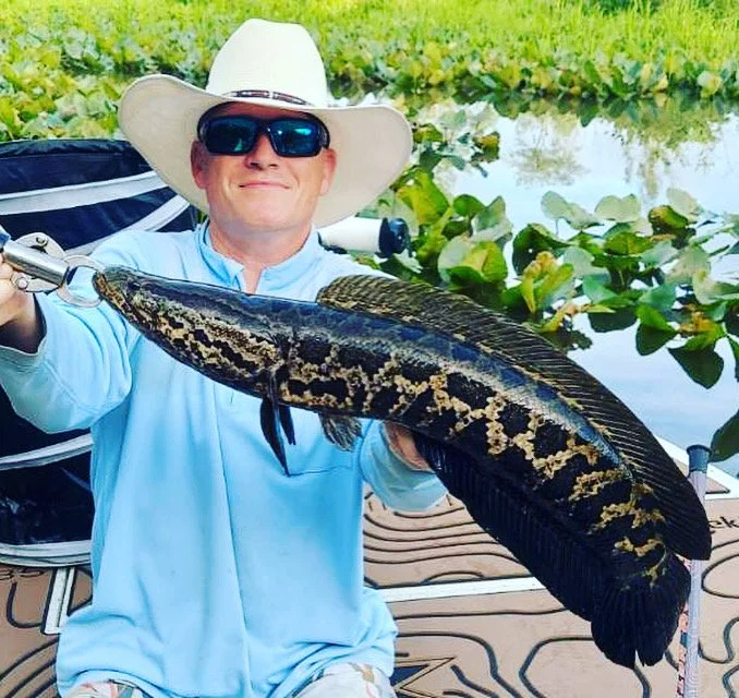 A man wearing a white cowboy hat and sunglasses, smiling while holding a large fish on a fishing line, with water and lily pads in the background.