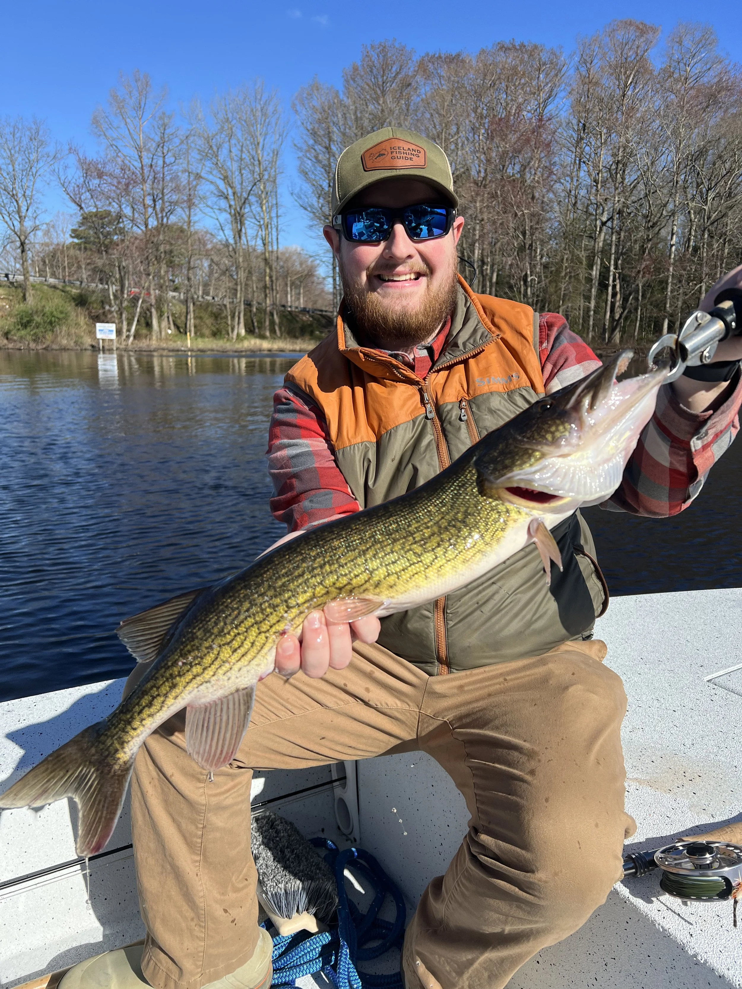 A man sitting on a boat holding a large fish, smiling and wearing sunglasses, an orange and brown vest, a cap, and khaki pants, with a body of water and trees in the background.