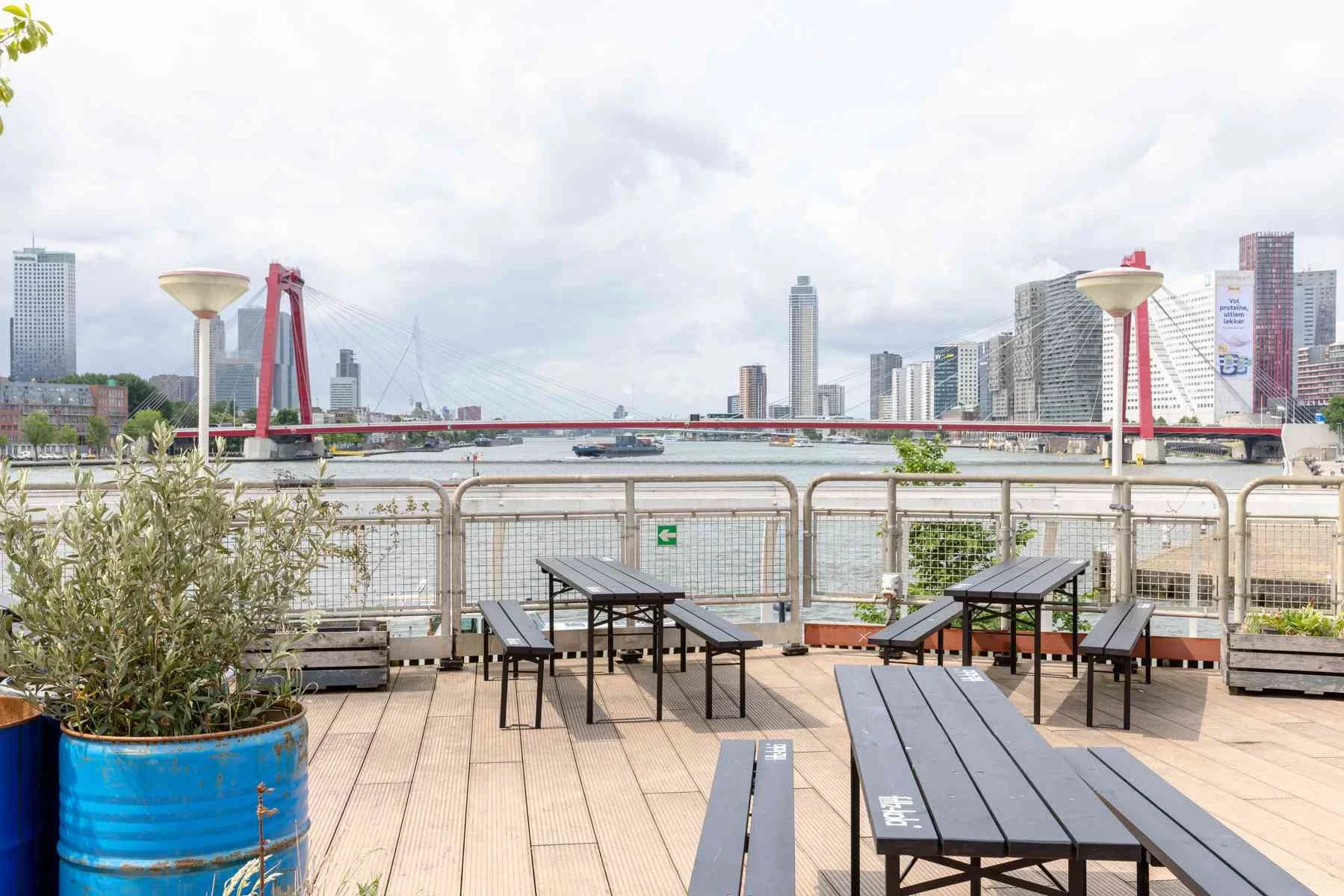 Outdoor seating area with wooden tables and benches, potted plants in a blue barrel, a metal railing, and a view of a river with a red bridge and city skyscrapers in the background under cloudy sky.