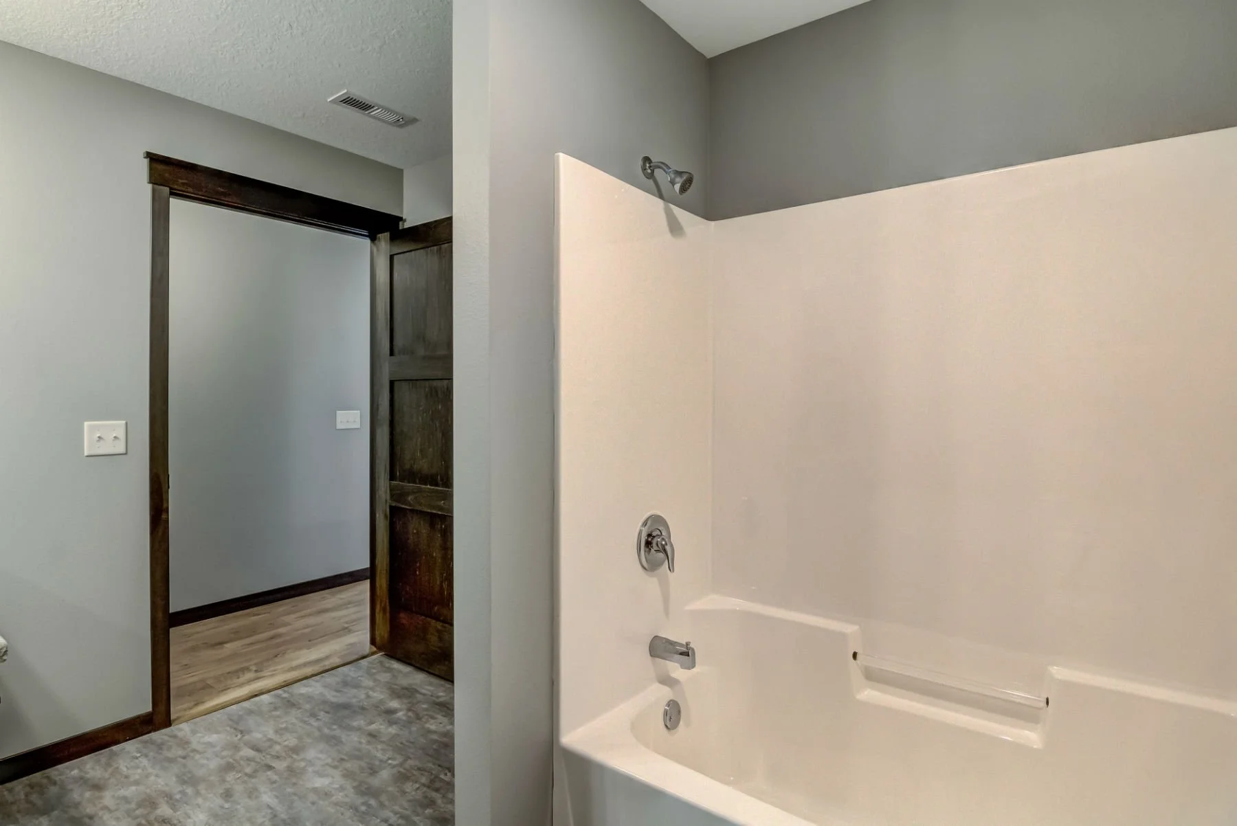 Empty bathroom with a bathtub, showerhead, and faucet, next to a doorway with dark wood trim leading to another room with wood flooring.