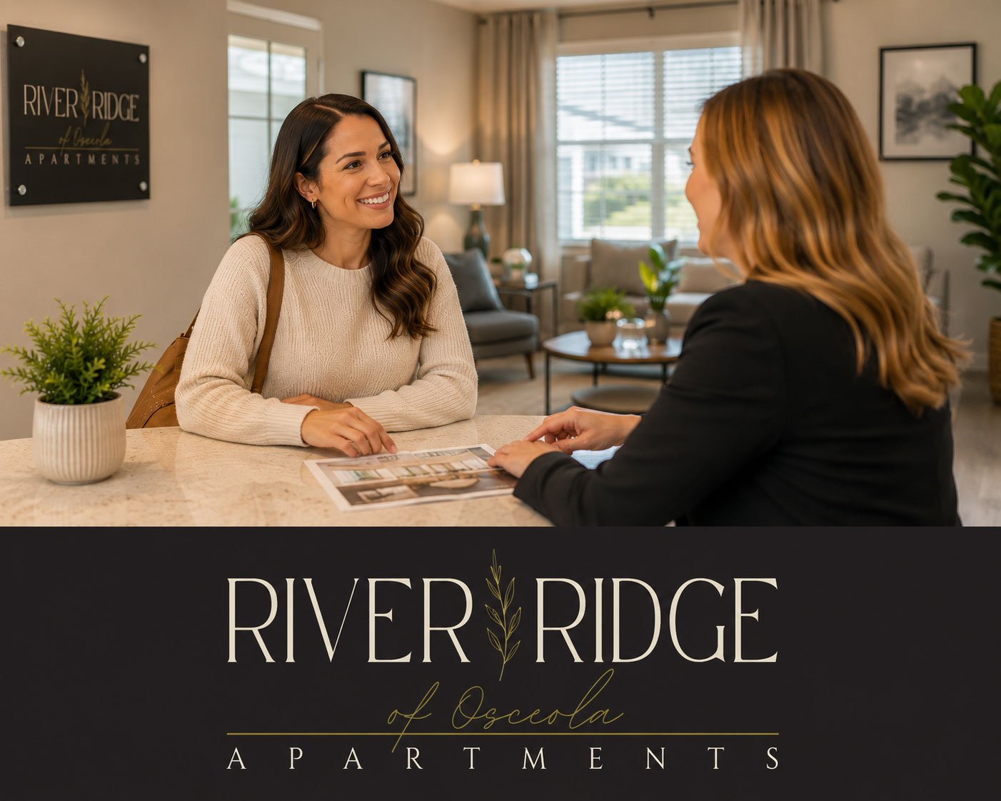 A woman with dark hair smiling and talking to a woman with reddish-blonde hair at a reception desk in a modern apartment leasing office. The office has large windows, a sofa, and plants. A sign on the wall reads 'River Ridge of Oscela Apartments.'