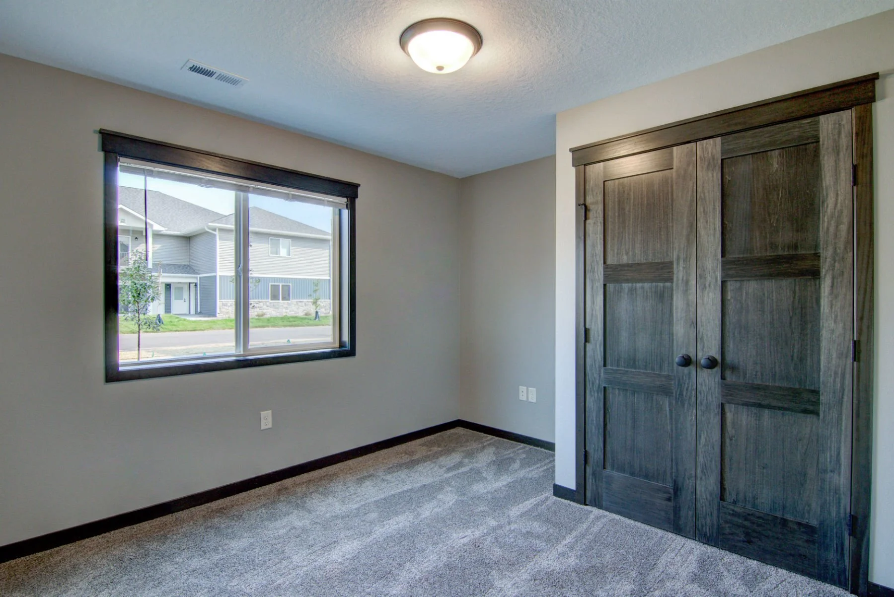 Empty bedroom with beige walls, a window revealing an exterior residential scene, a ceiling light fixture, and dark wood closet doors.