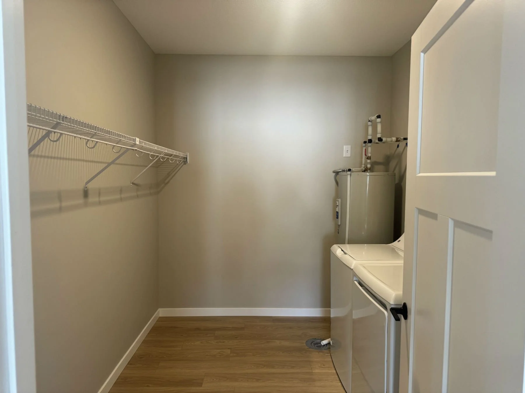 Small laundry room with beige walls, wood-look flooring, a white washing machine, a water heater in the corner, a wall outlet, a drain in the floor, and a wire shelf on the left wall.