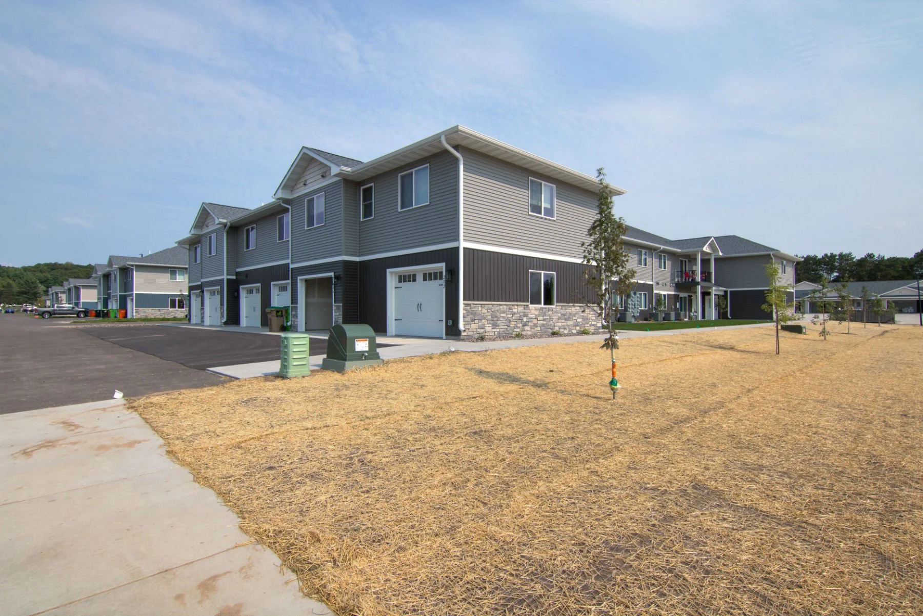 A row of new modern townhouses with gray siding, white trim, and stone accents, in a residential area with young trees and a partly paved yard under a clear sky.